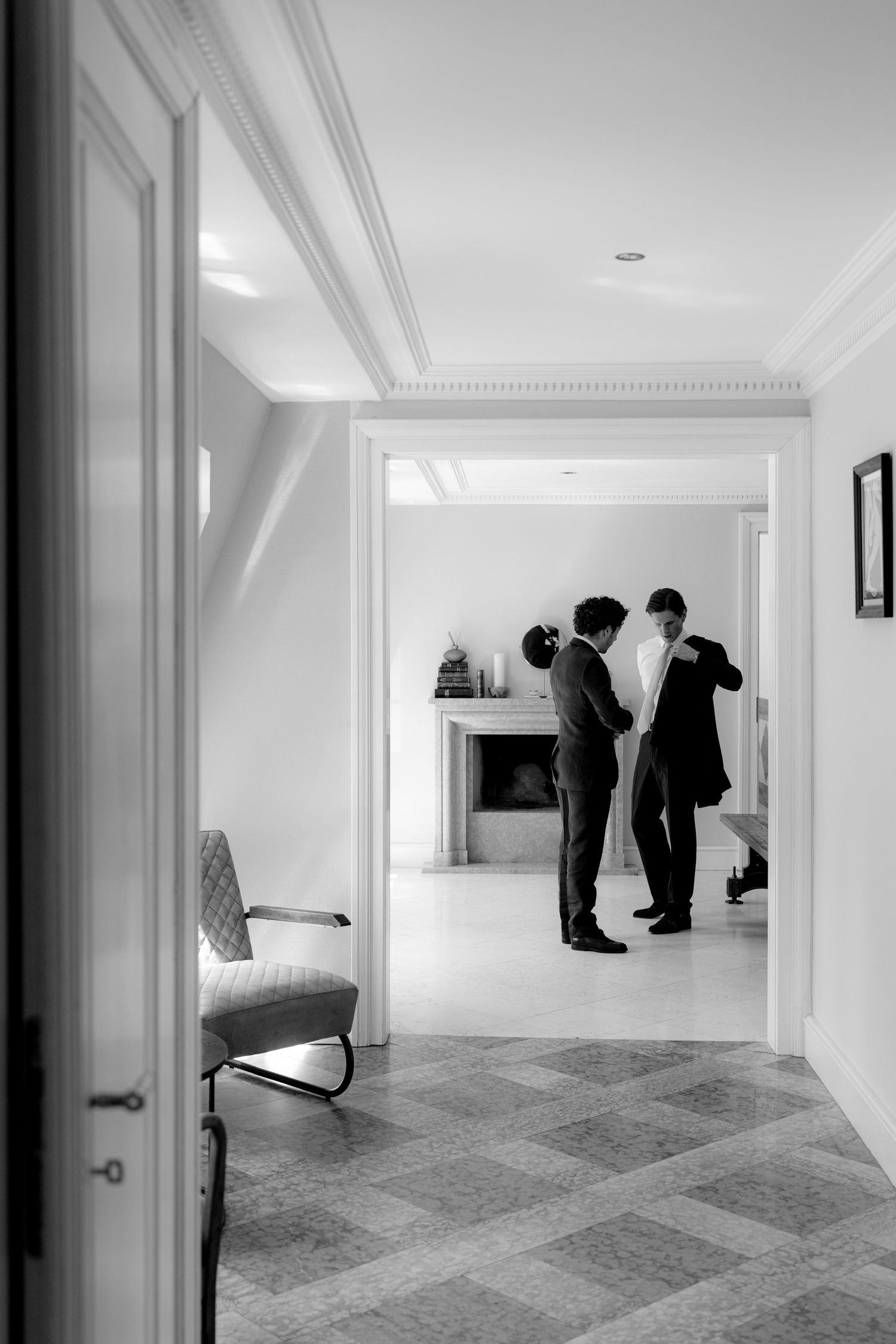 Black and white photo of two men in suits standing in an elegant hallway with fireplace and classic chair.
