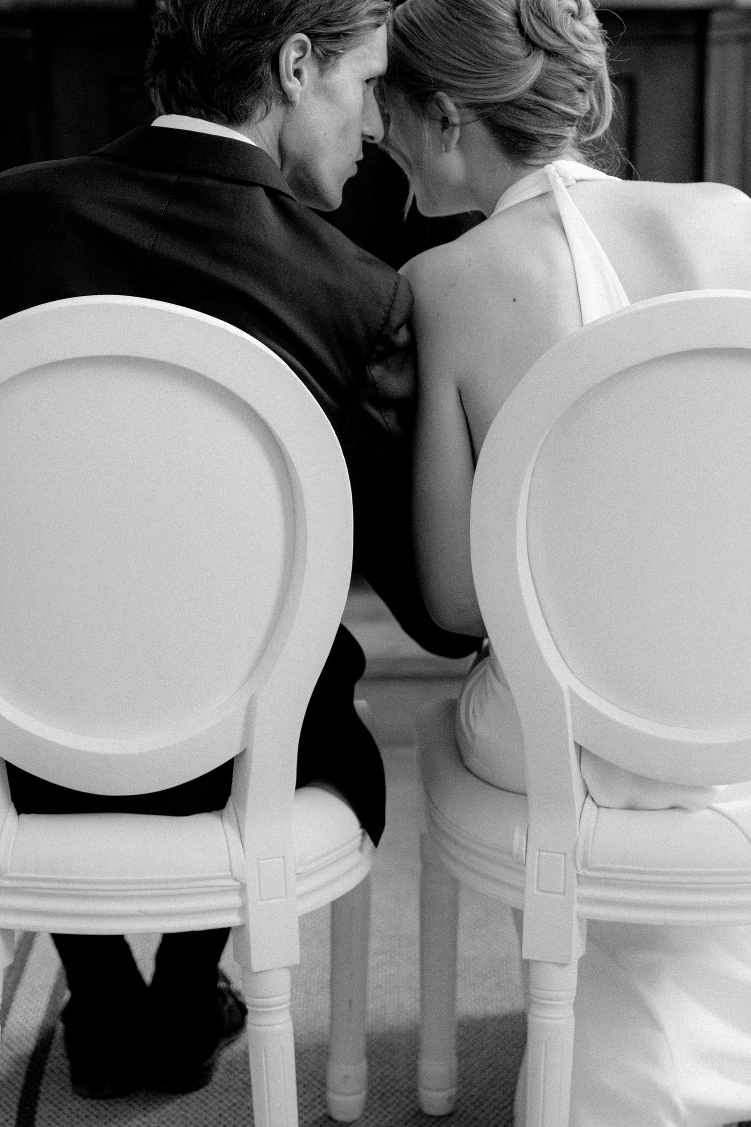 Black and white photo of bride and groom sitting in elegant white chairs, leaning close together at their wedding ceremony.