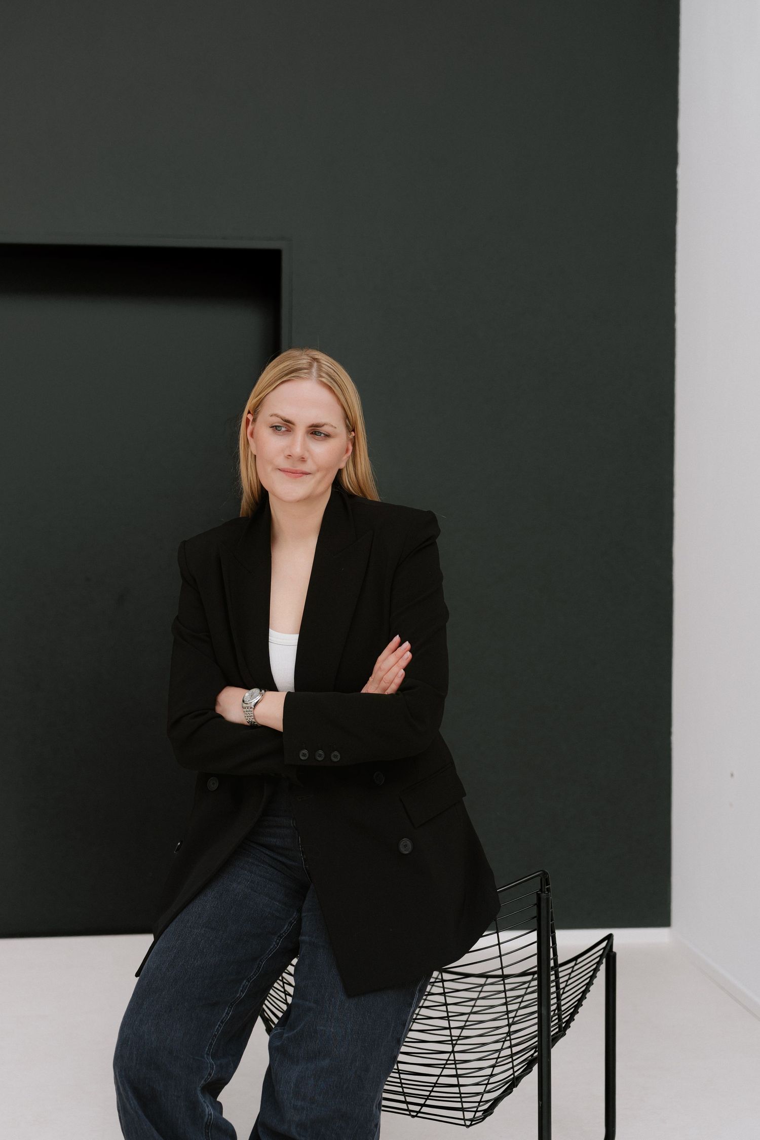 Blonde woman in black blazer and jeans poses with arms crossed against a dark modern wall.