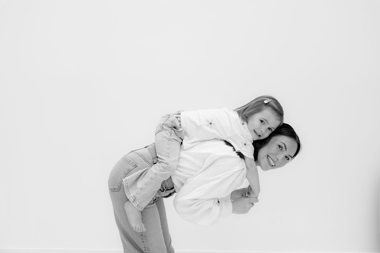 Black and white photo of a woman giving a smiling girl a piggyback ride against a white background.