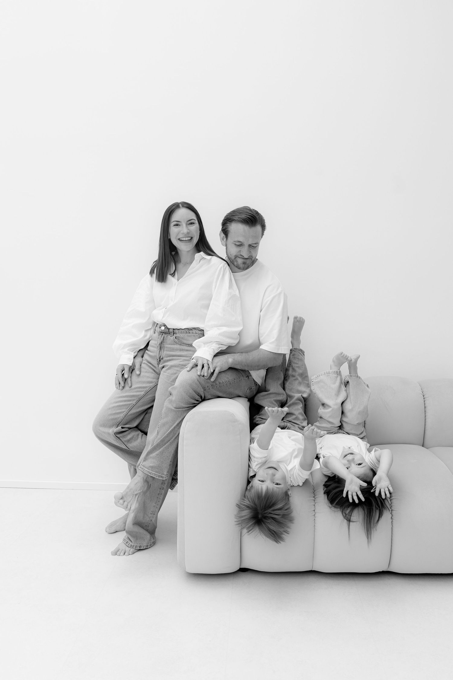 Black and white family photo with parents and two children playing on a modern white sofa in a bright studio.