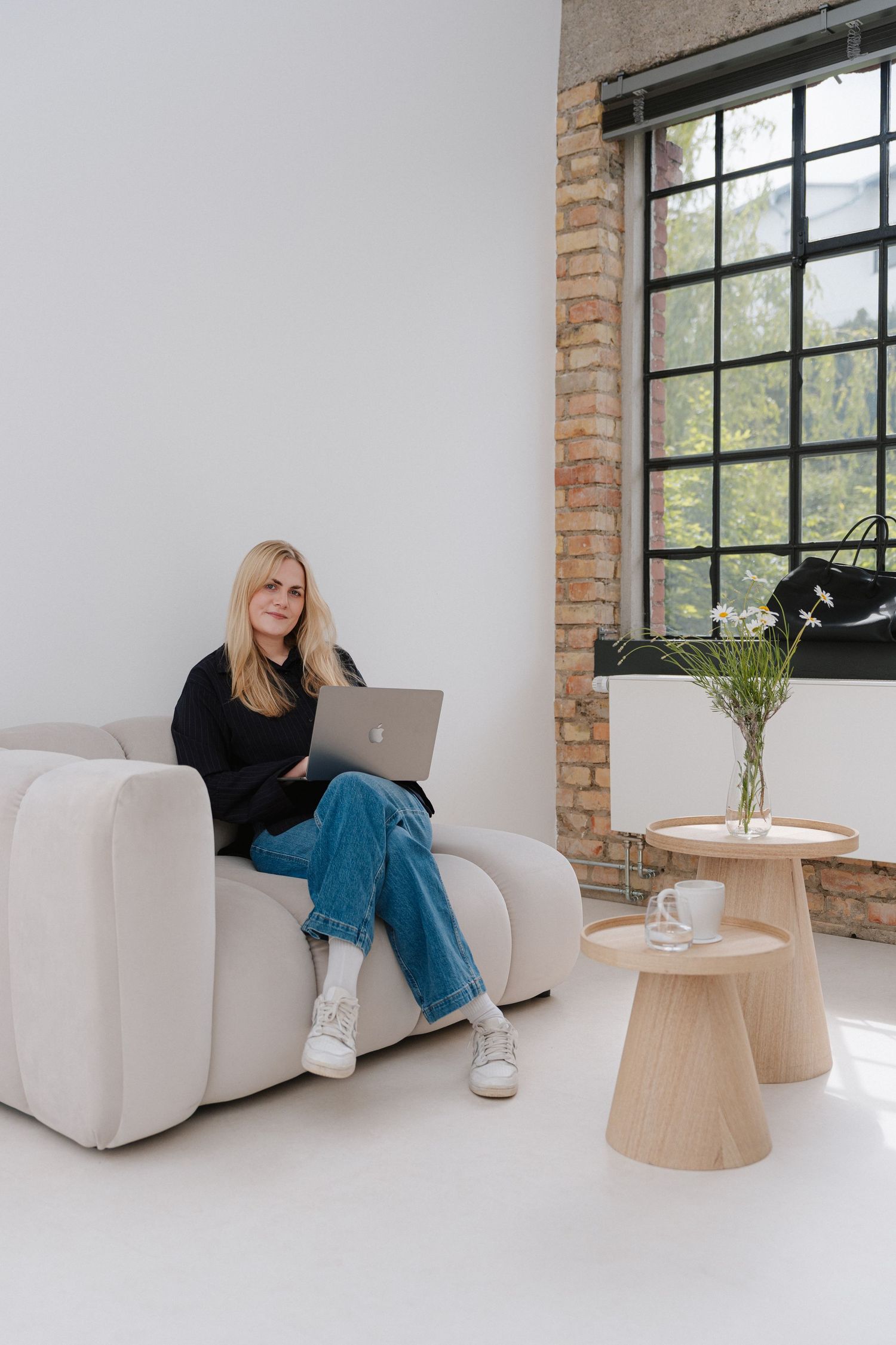 Woman with blonde hair sitting on white sofa with laptop in modern loft space with brick wall and large windows.
