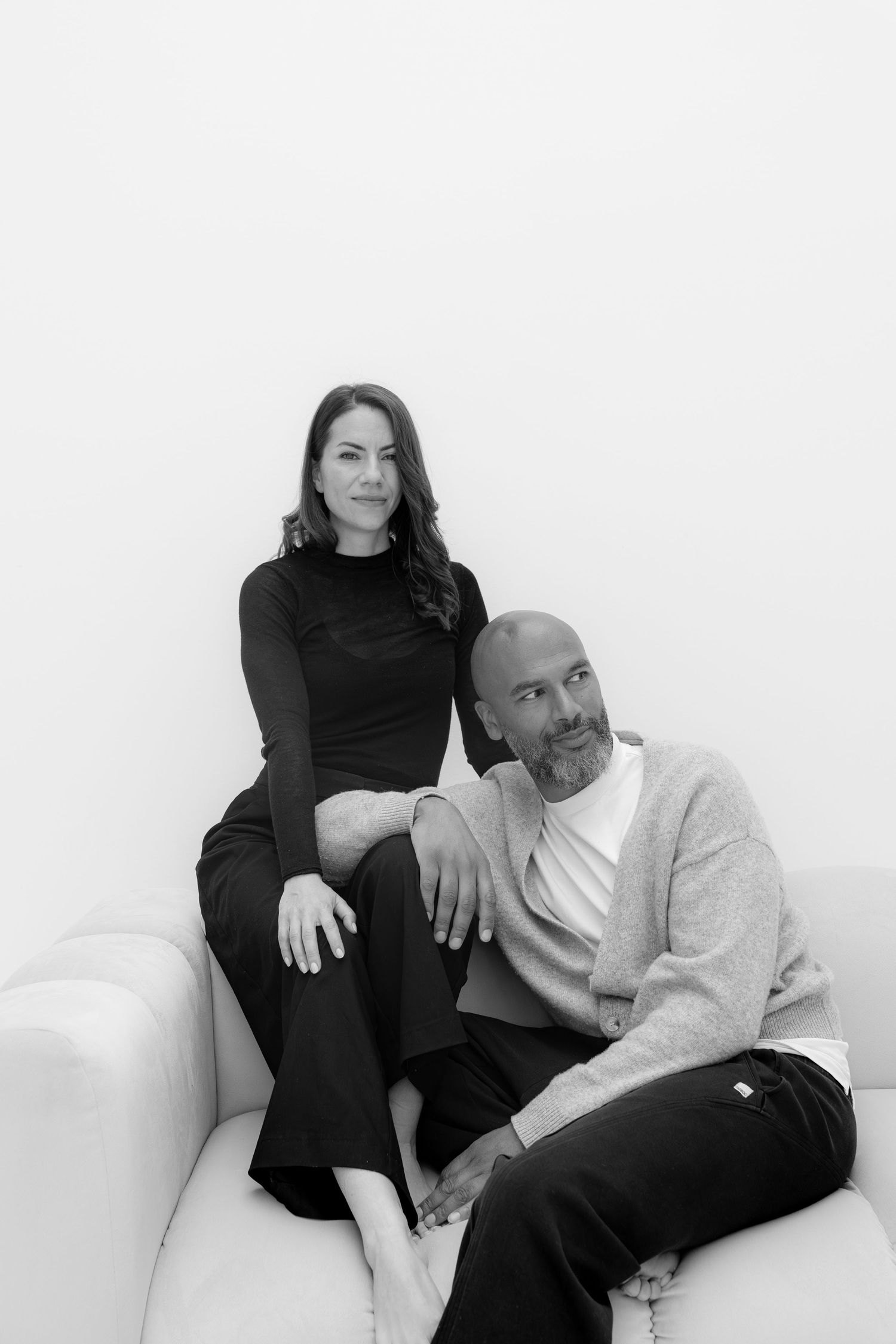Black and white portrait of a woman and bald man with beard sitting together on a white sofa.