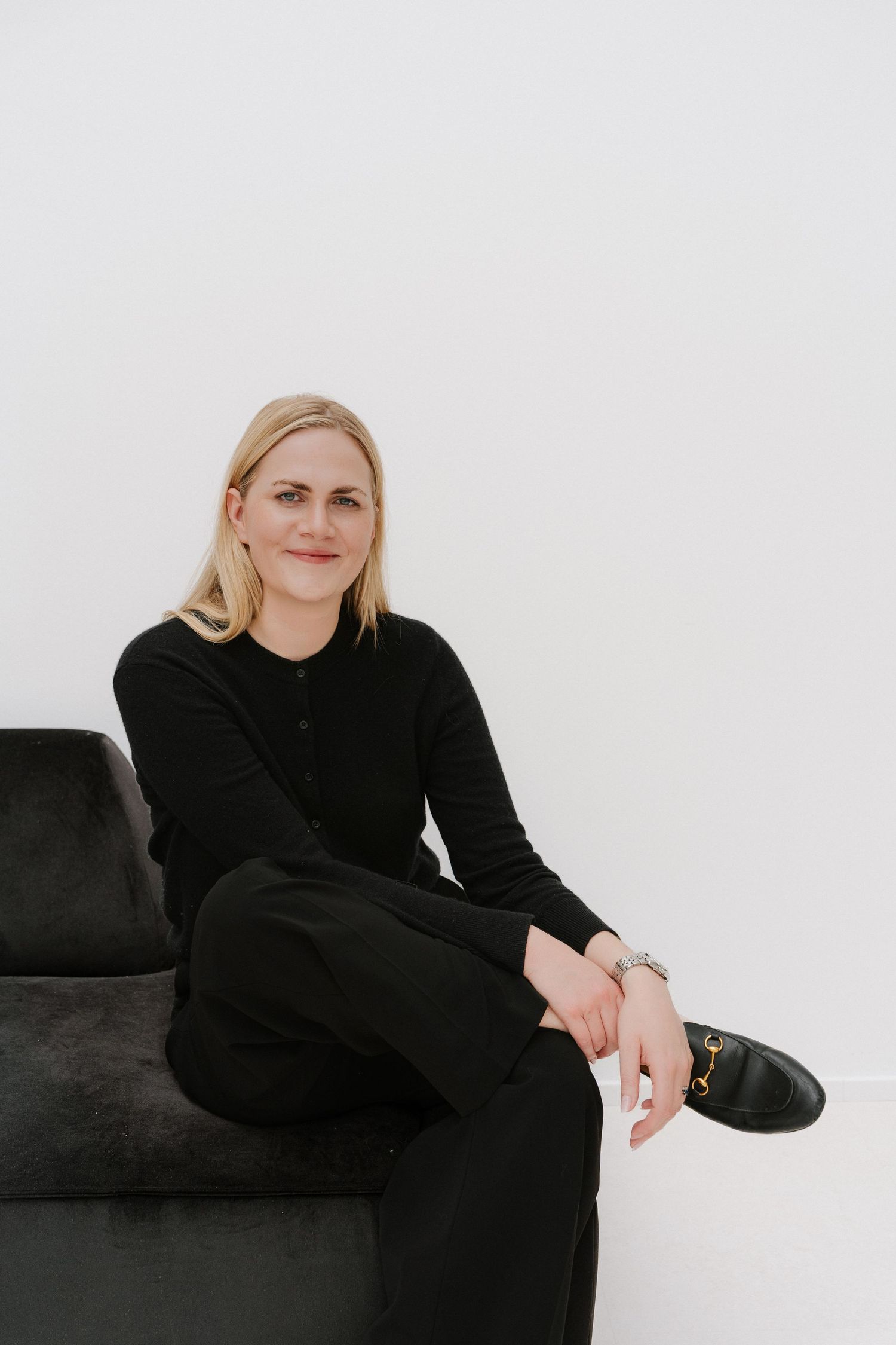 Blonde woman in black outfit sitting on dark velvet sofa against white background, smiling confidently.