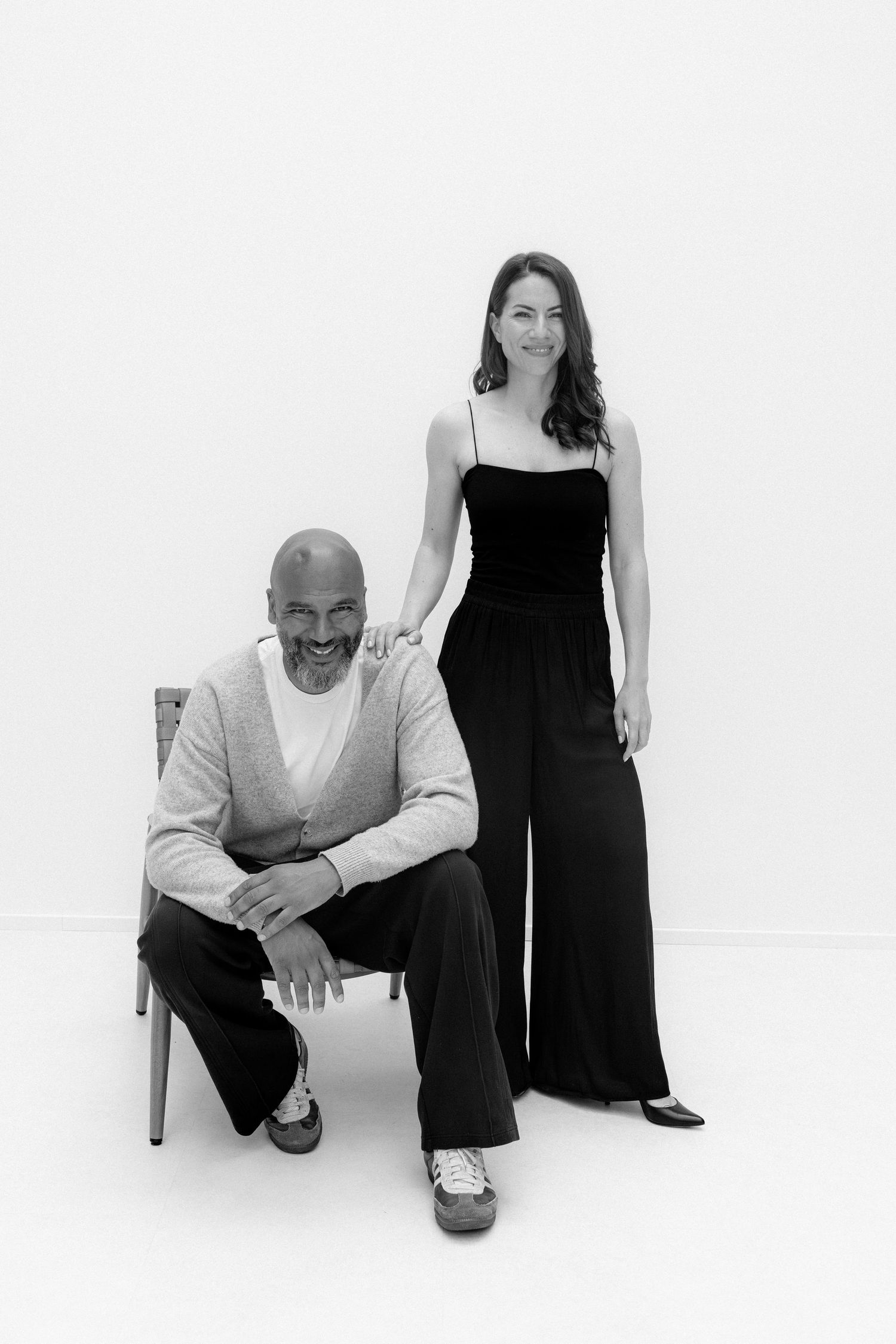 Black and white portrait of a bald man seated and a woman standing beside him, both smiling in elegant attire.
