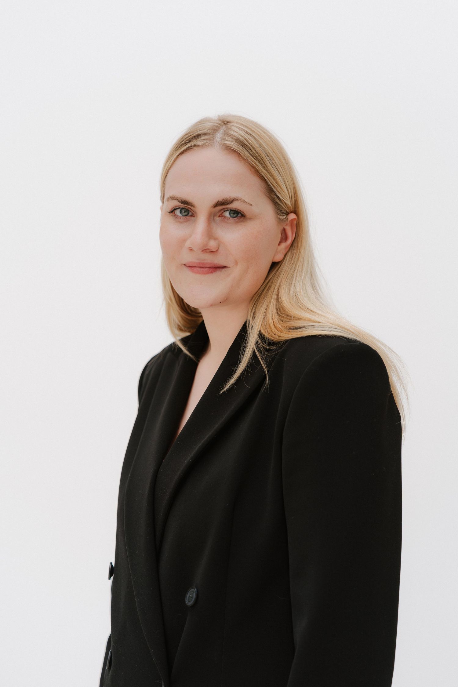 Professional blonde woman in black blazer poses against white background for corporate headshot.
