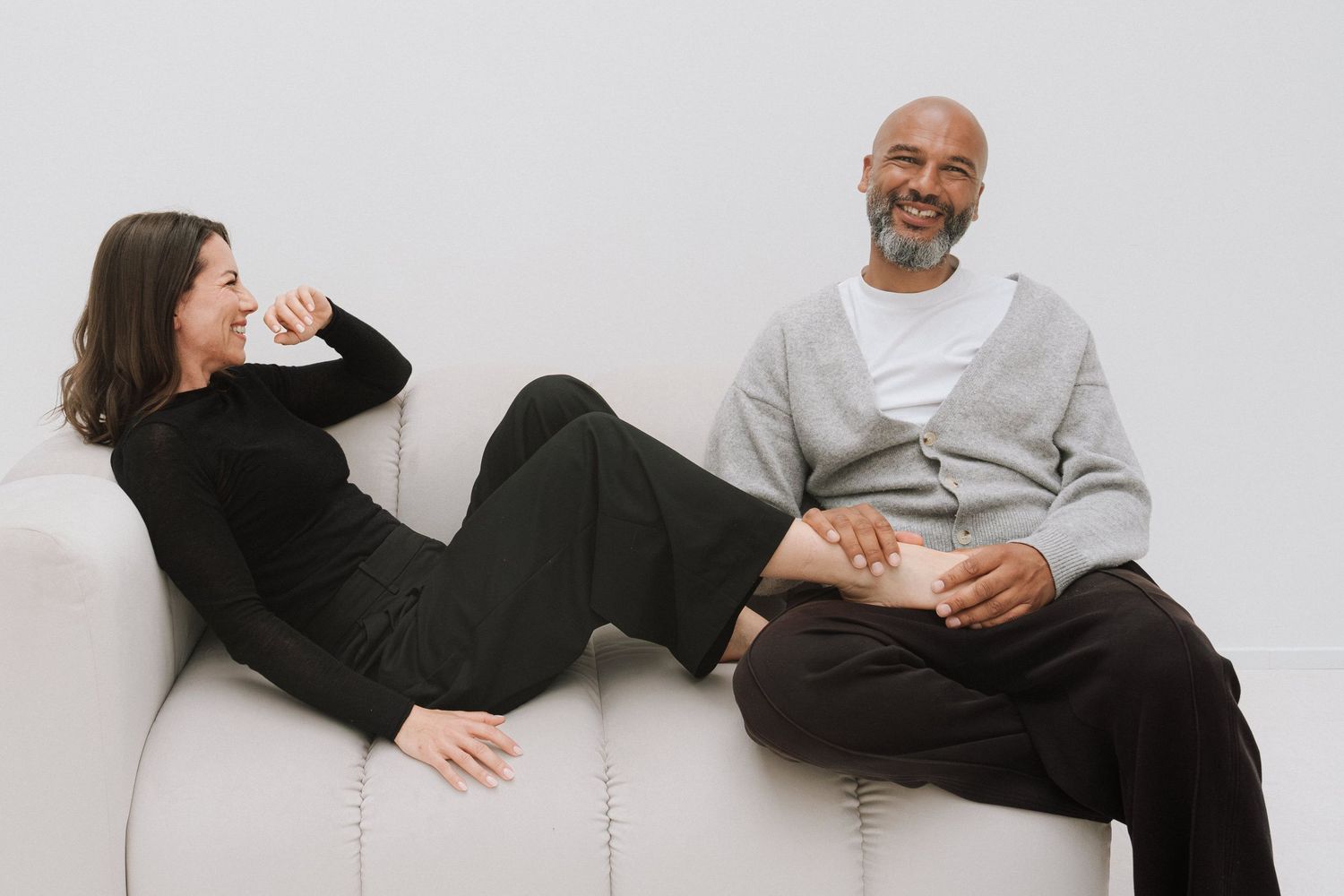 A man and woman laughing together on a white couch in a bright, minimalist setting.