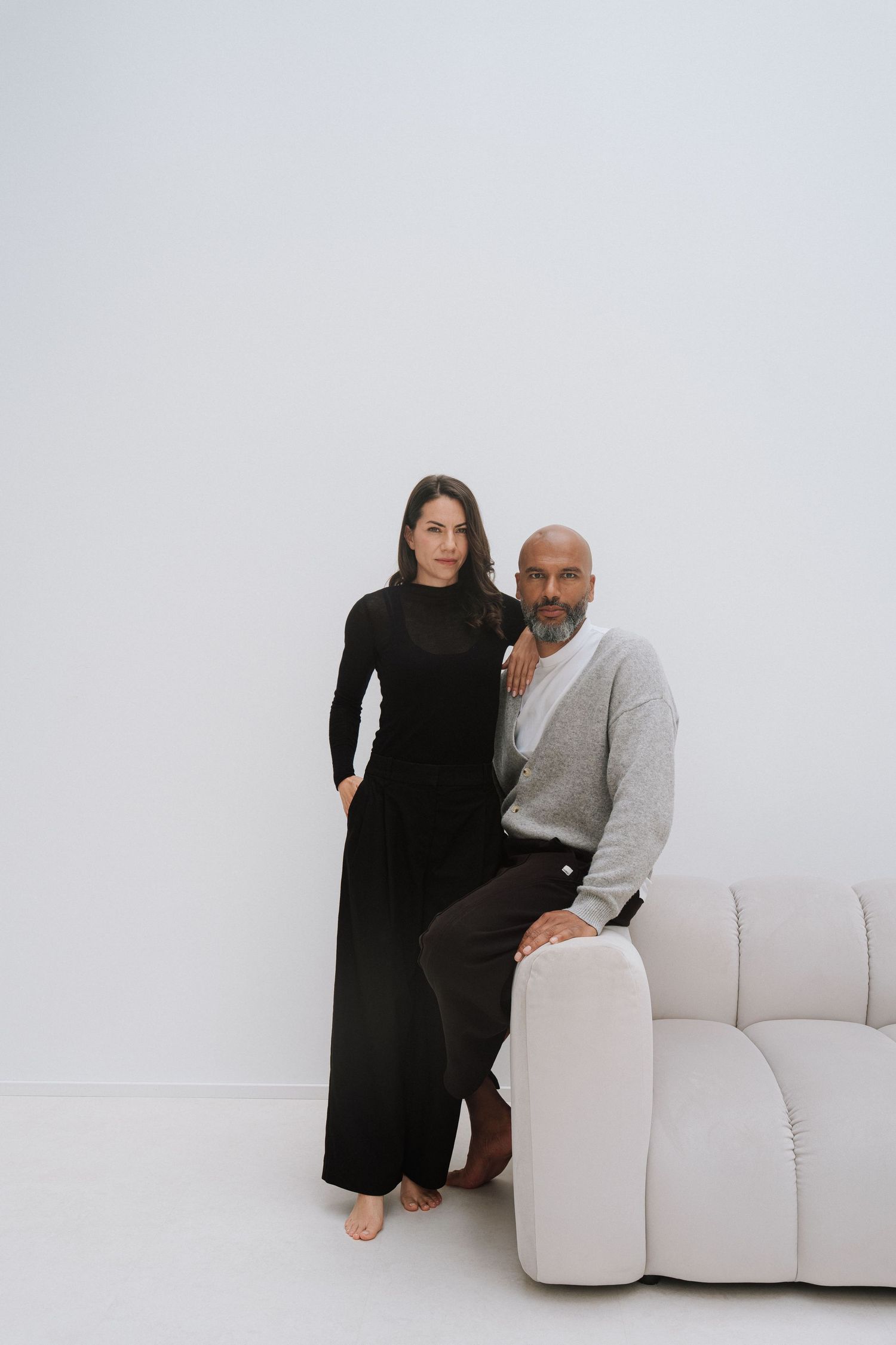 A man and woman pose elegantly in a minimalist white studio, she in a black dress, he in a gray sweater near a cream sofa.