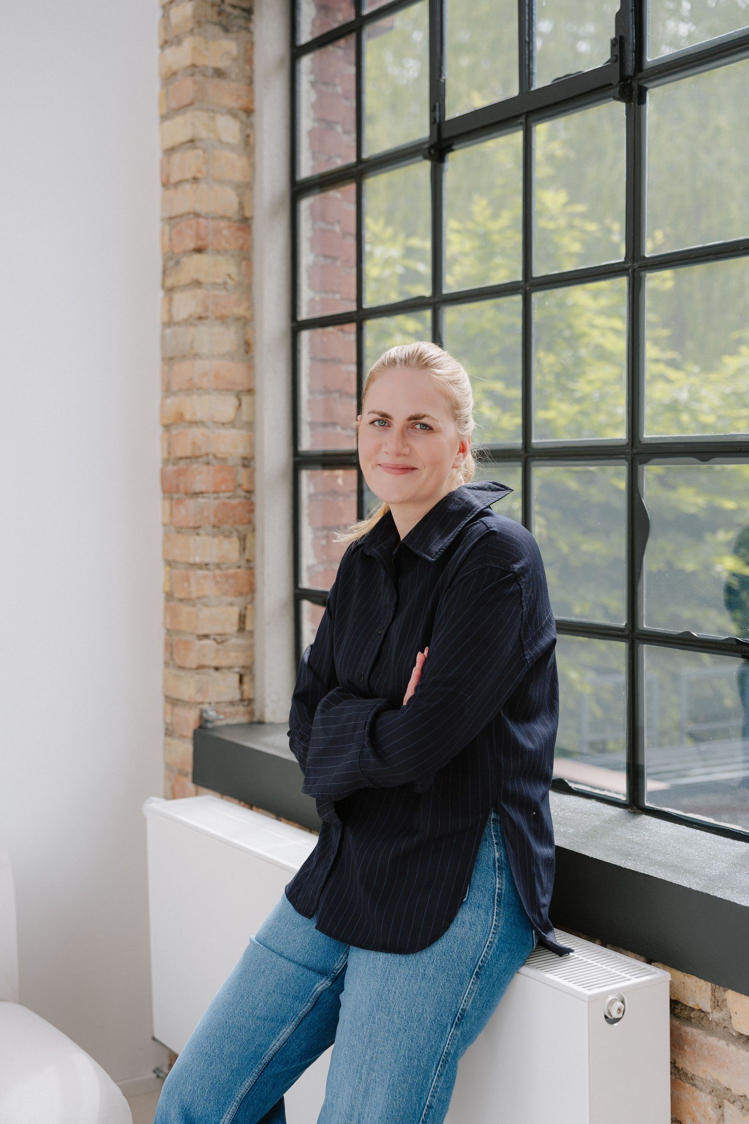 Woman in dark shirt and jeans leaning against windowsill in industrial loft with brick walls and large windows.