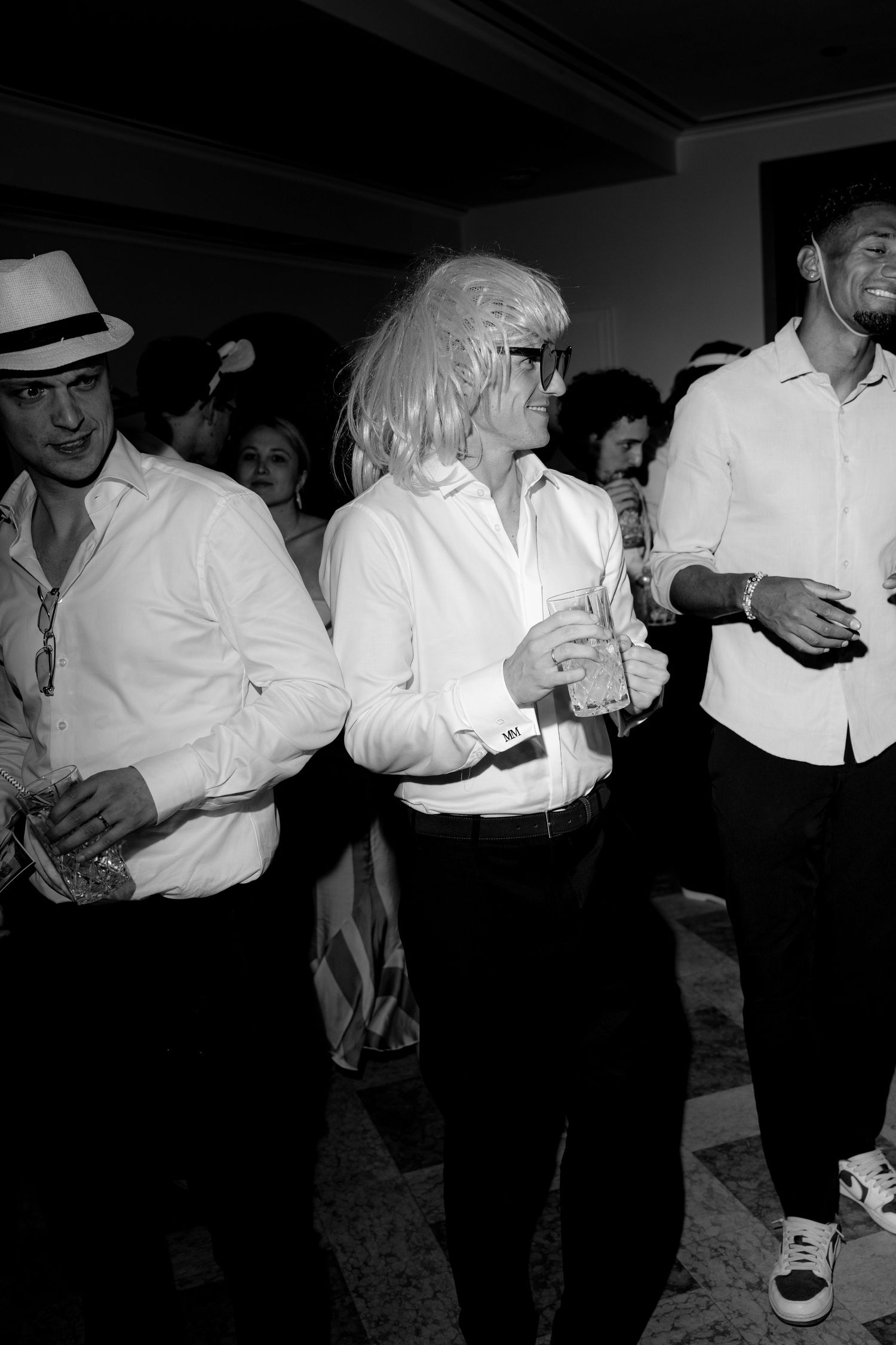 Black and white photo of party guests in white shirts, one wearing a blonde wig and glasses, holding drinks.