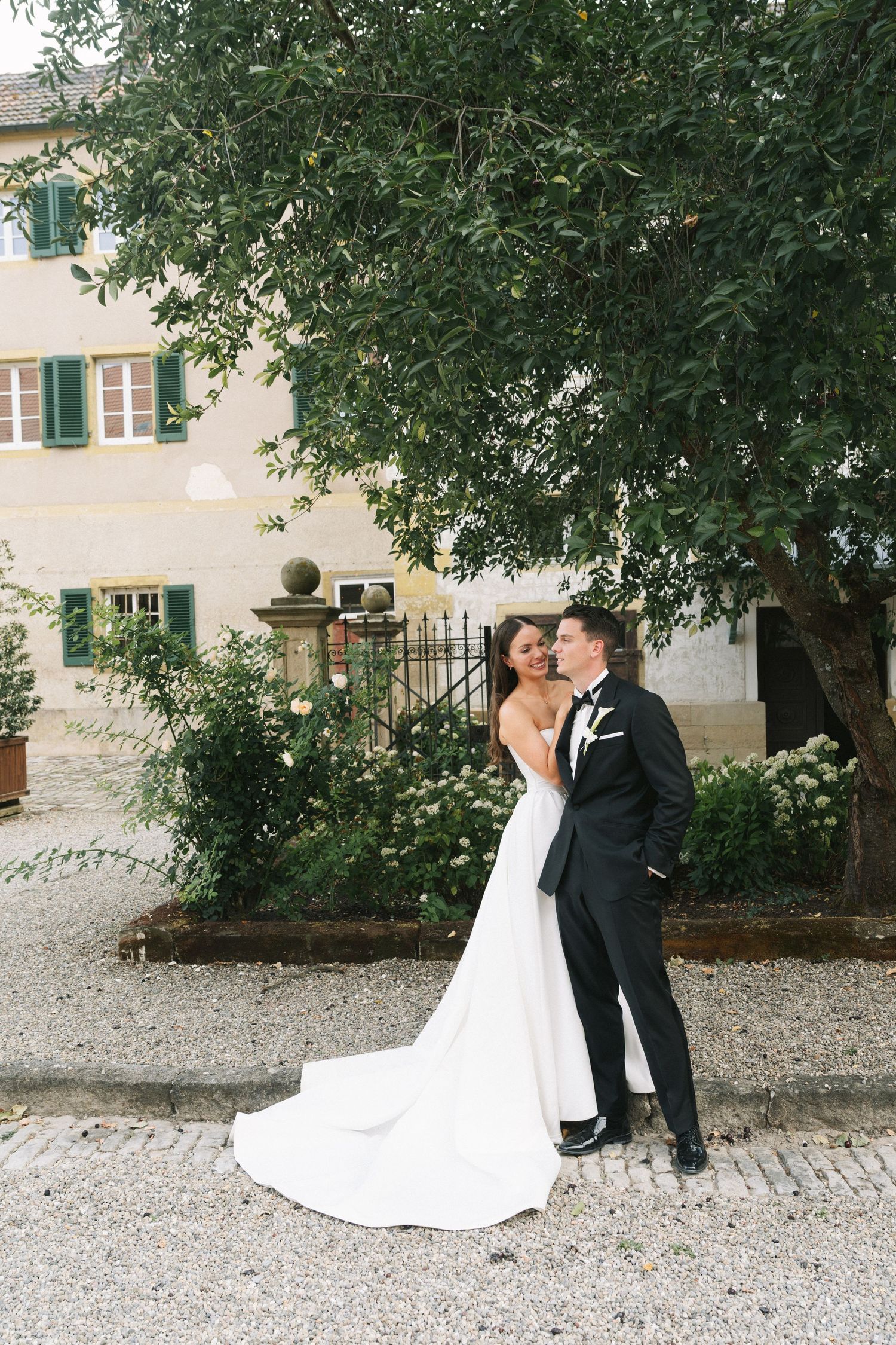 Bride and groom embrace near a lush tree outside an elegant European villa on their wedding day.