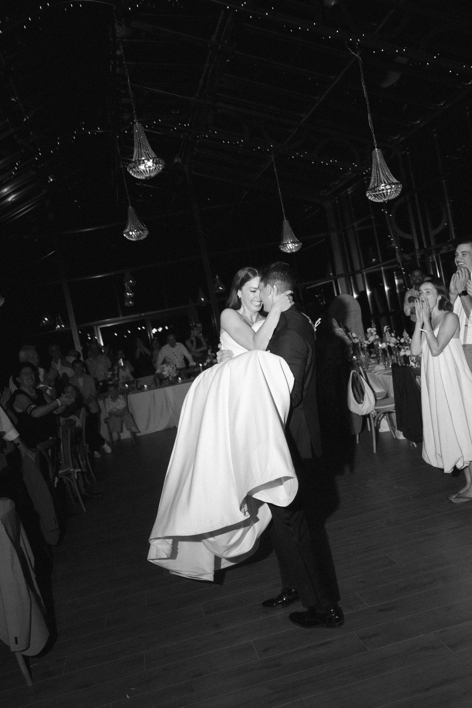 Black and white photo of groom lifting bride during first dance at elegant wedding reception with chandeliers.