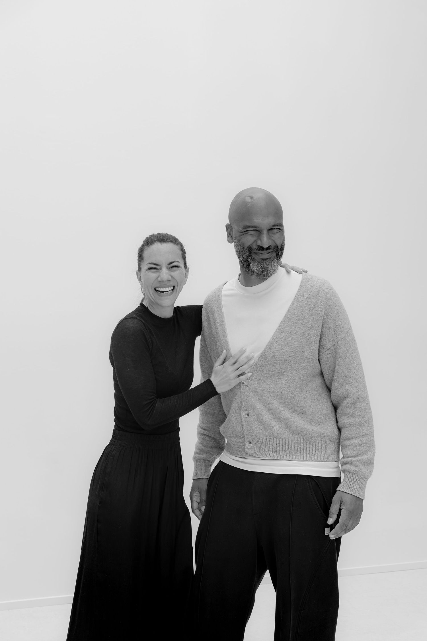 Black and white photo of a smiling woman in black dress and bald bearded man in cardigan posing together.