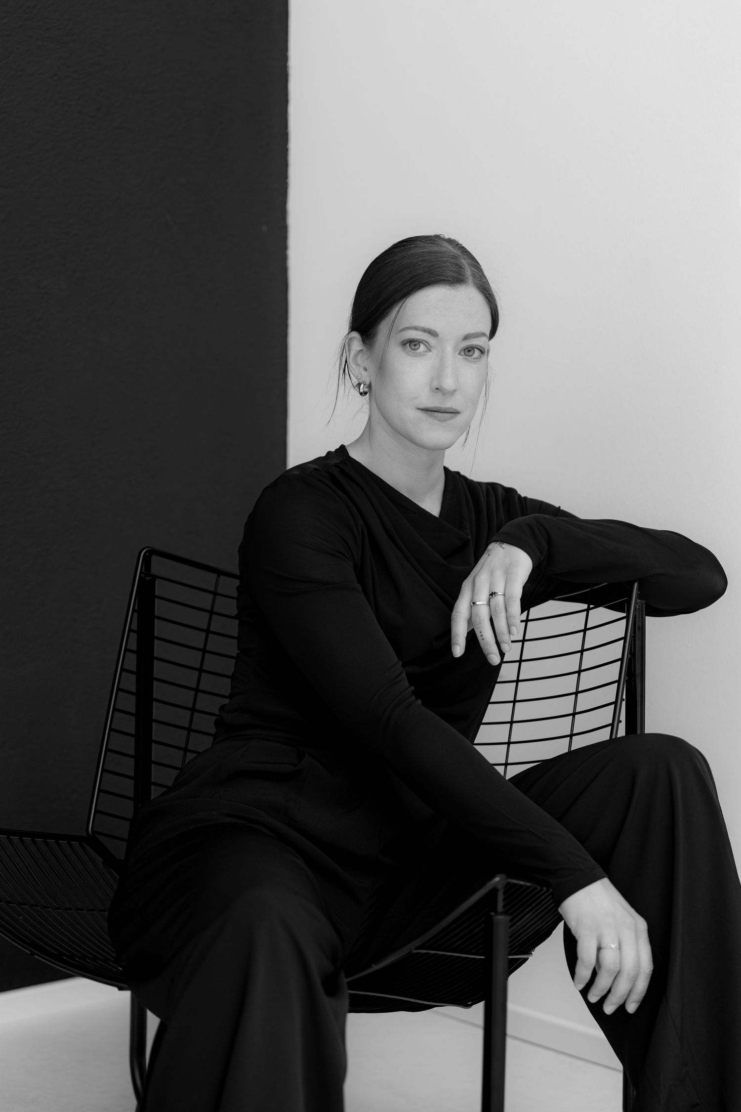 Black and white portrait of a woman in black outfit seated on a metal chair against a two-toned wall.