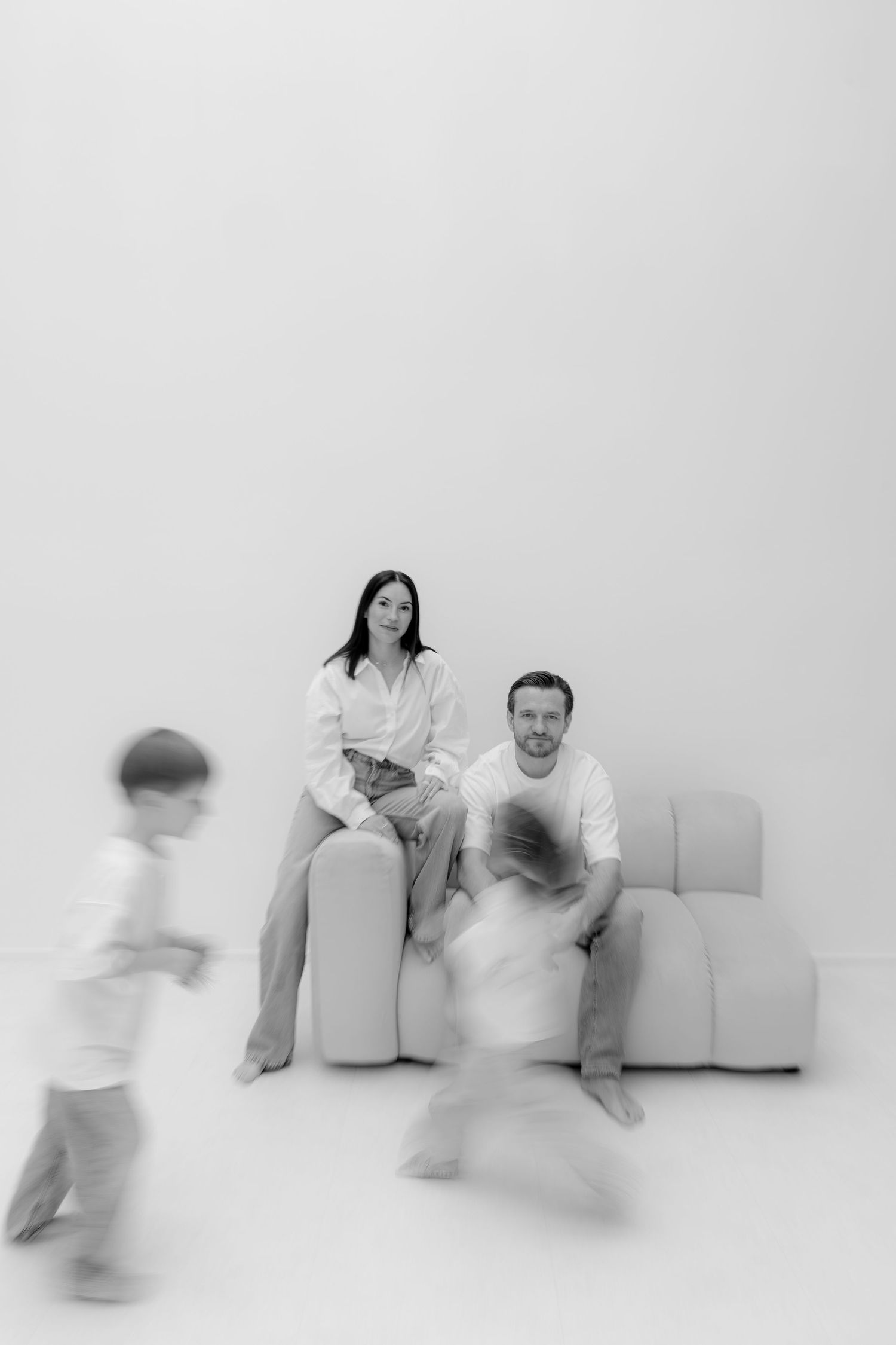 Black and white family photo of parents on sofa while two children run and play in a bright white room.