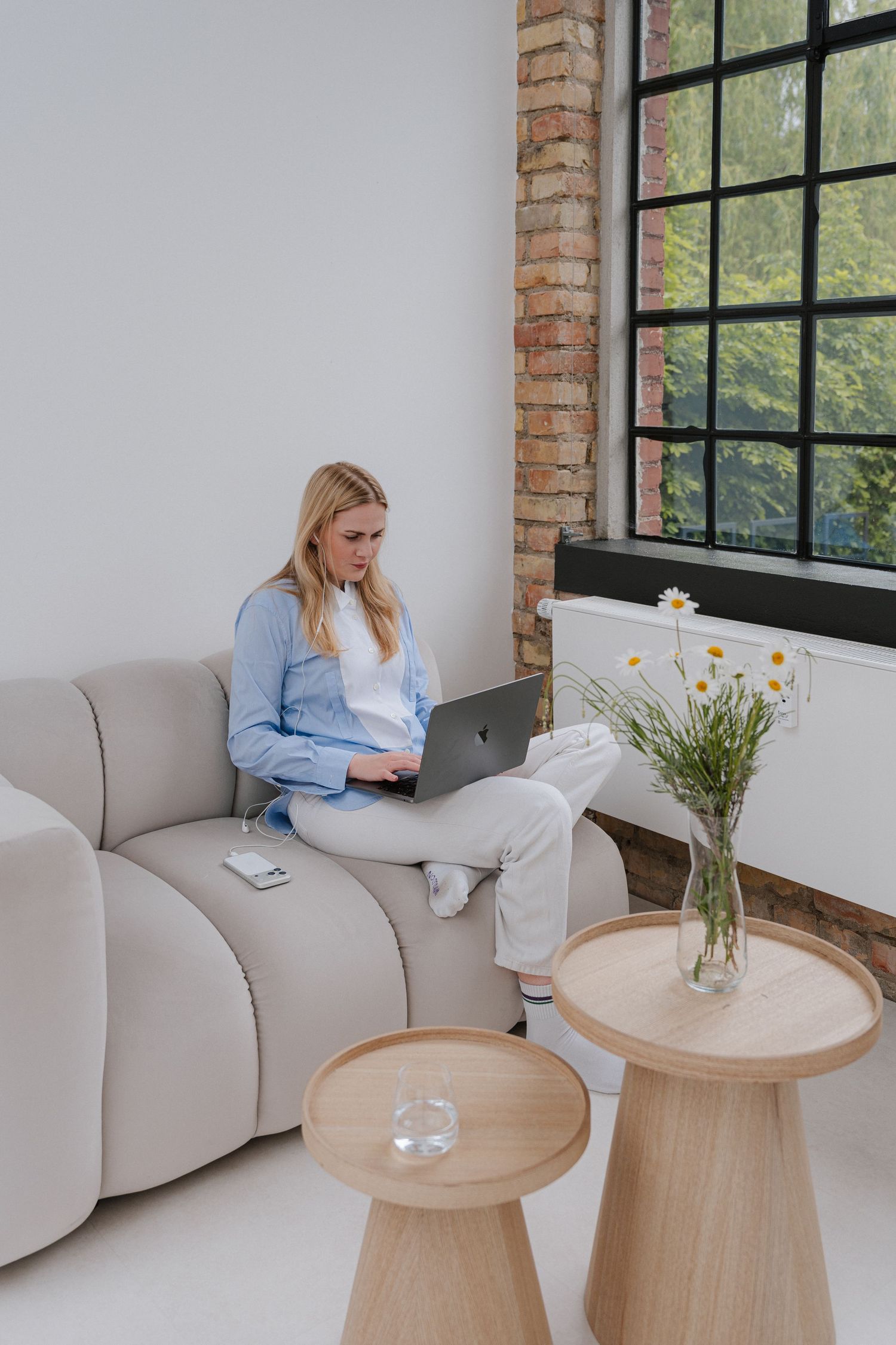 Woman working on laptop while sitting on modern gray sofa near large window with wooden side tables and flowers.