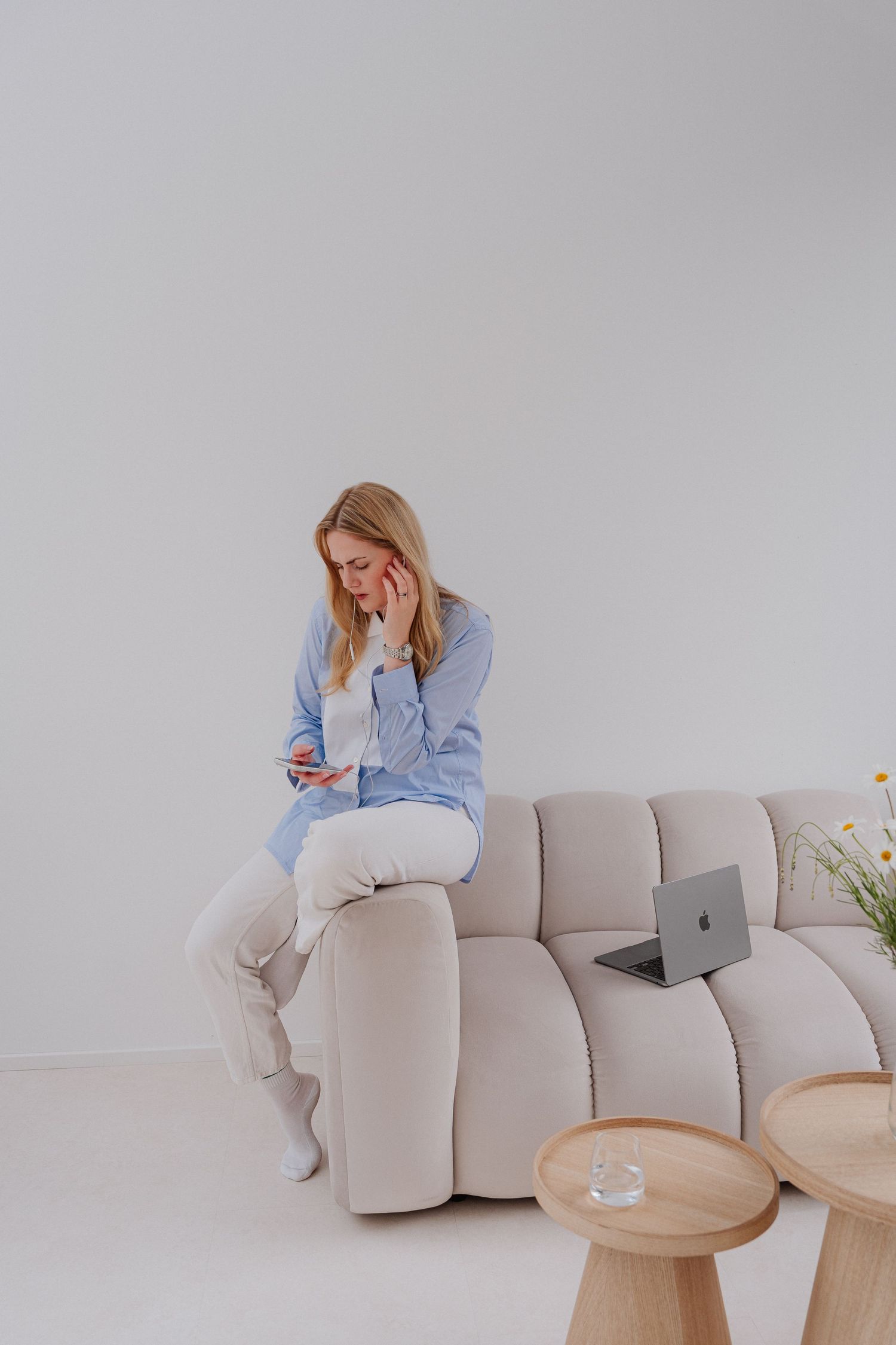 Woman in blue shirt sitting on beige sofa using smartphone with laptop beside her in minimalist white room.