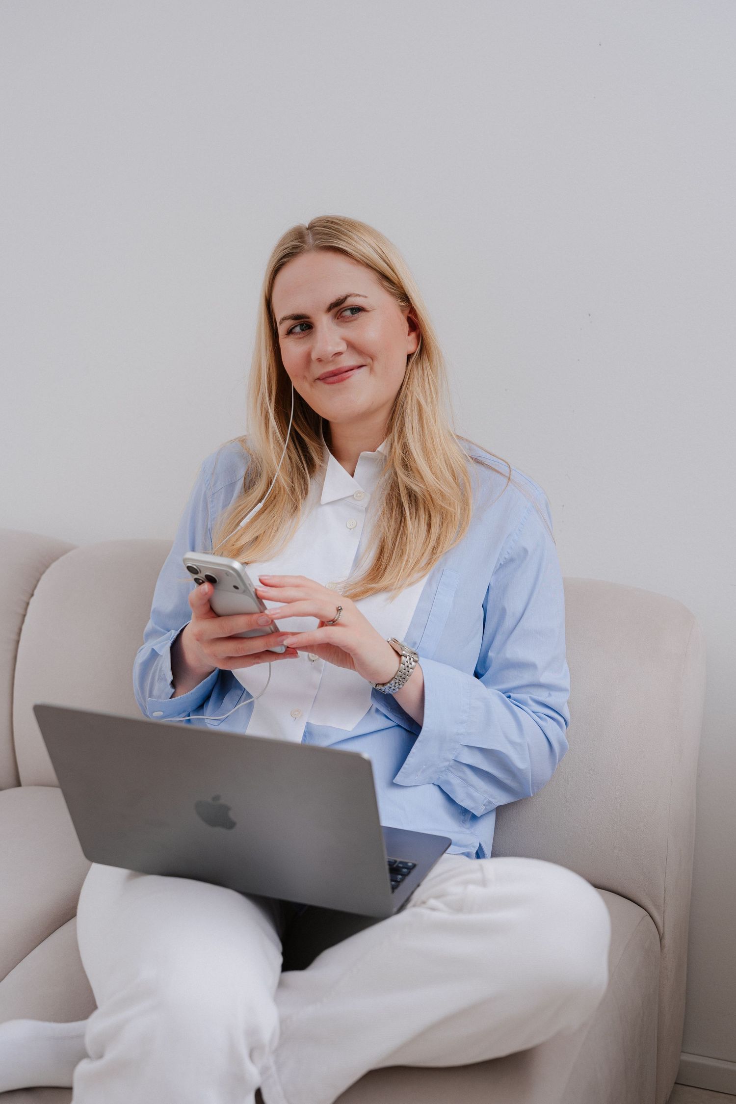 Woman sitting on sofa with laptop and smartphone, wearing blue blazer and white pants, smiling at camera.