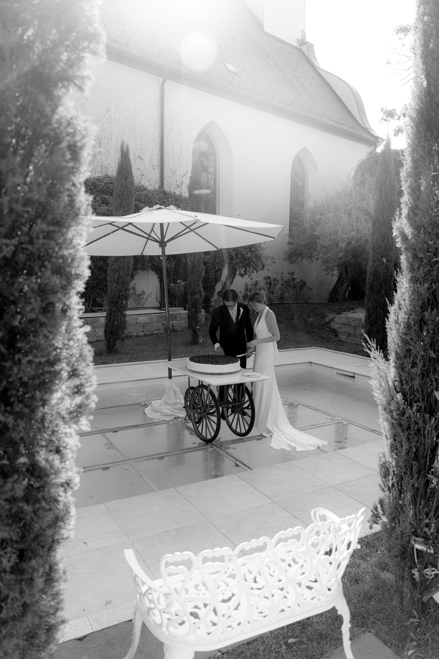 Black and white photo of bride and groom cutting wedding cake outdoors under umbrella beside elegant church.