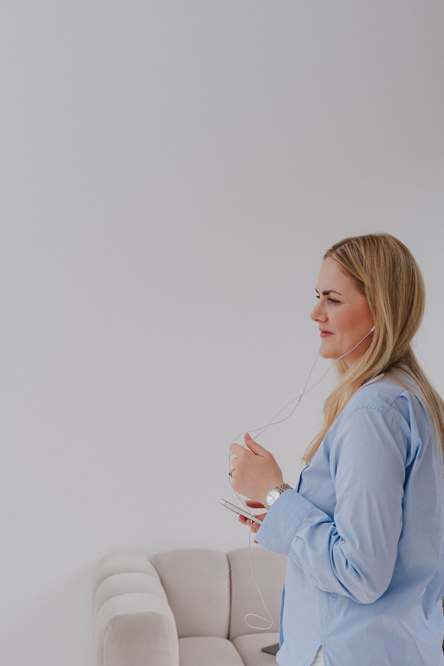 Blonde woman in blue shirt holding glasses, seated by a white sofa against a light gray background.