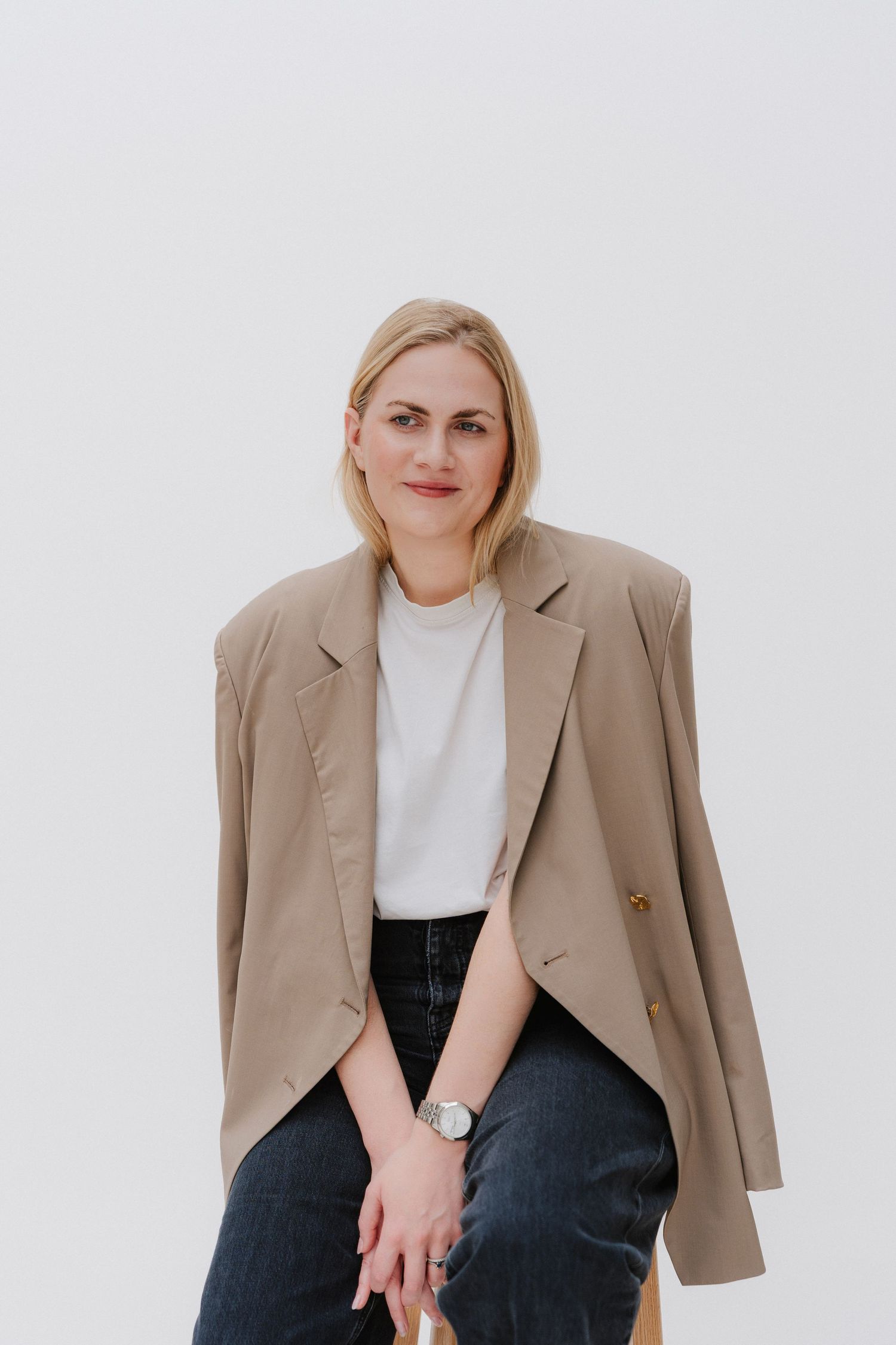 Blonde woman in beige blazer and white tee sits confidently against white background wearing a silver watch.