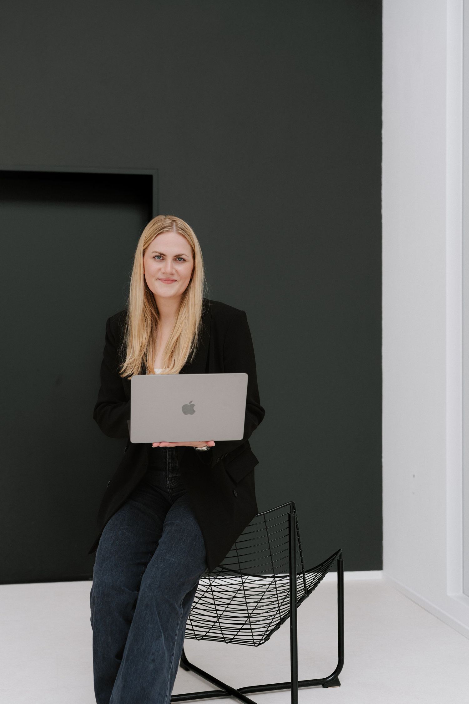 Blonde woman in black blazer sitting on wire chair holding MacBook laptop against dark green wall.
