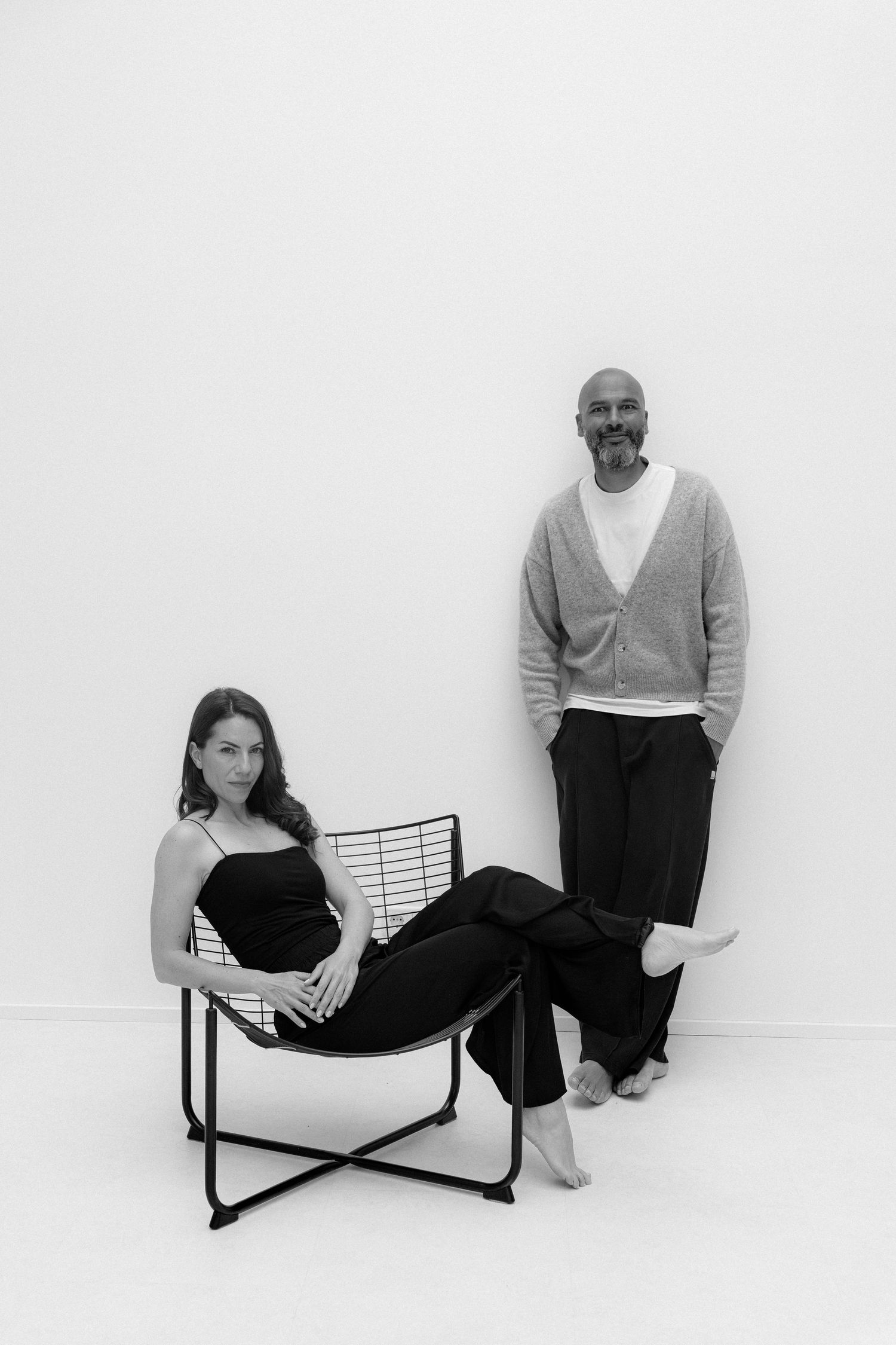 Black and white photo of a woman sitting in a wire chair and a bald man standing against a white wall.