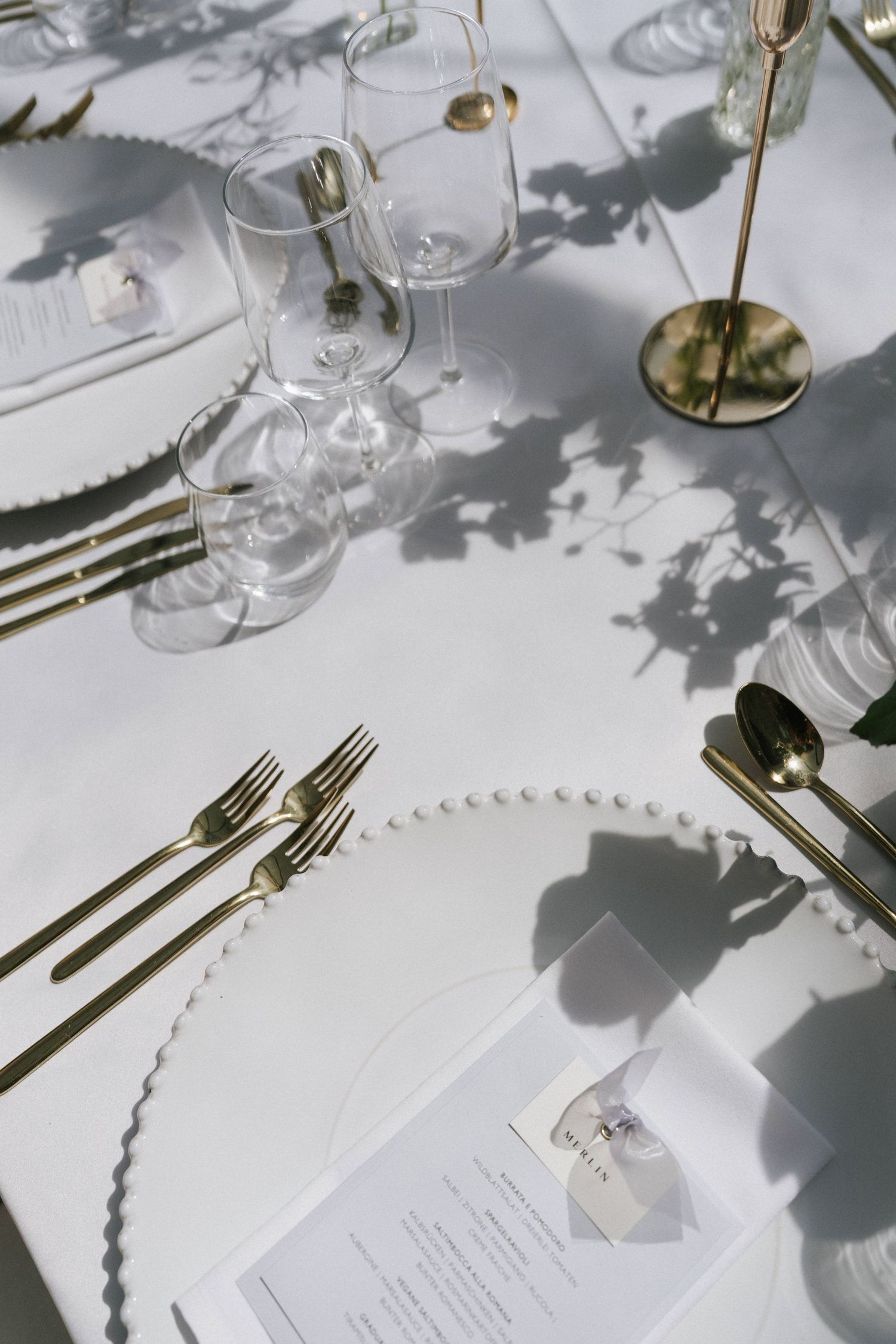 Elegant wedding table setting with gold cutlery, pearl-edged plates, crystal glasses, and floral shadows on white linen.