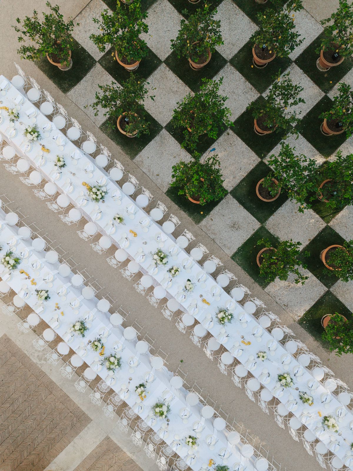 Aerial view of elegant outdoor wedding reception with long white-draped tables, chairs, and potted trees on a checkered stone patio.