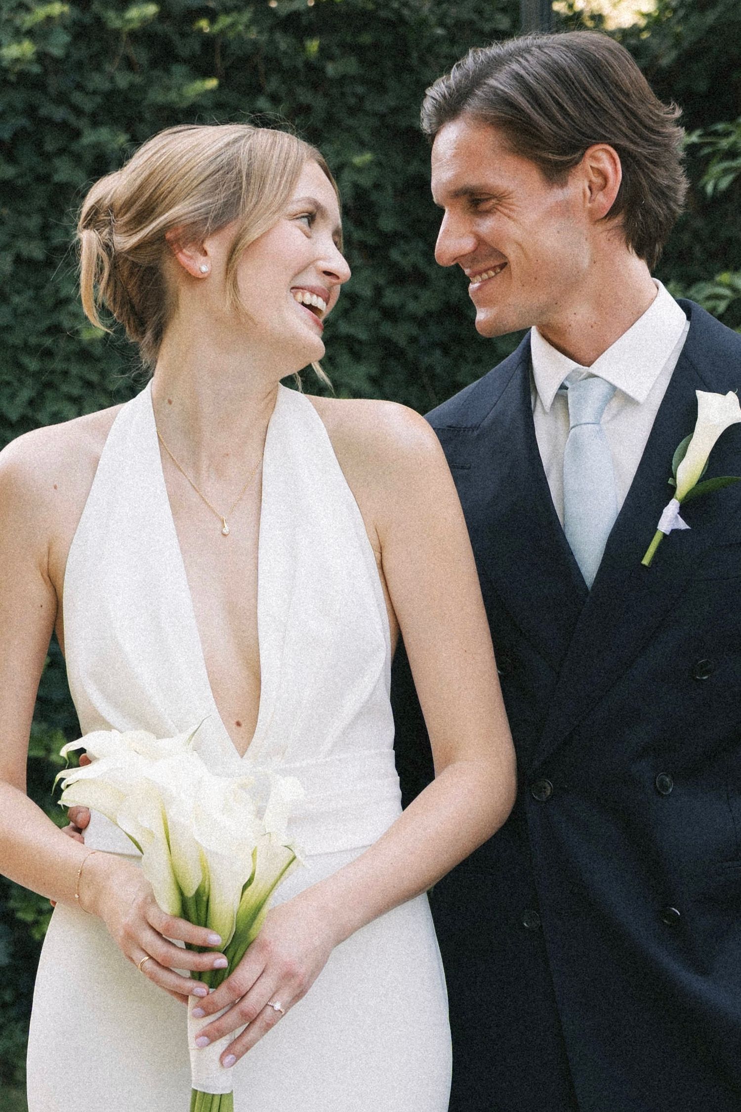 A smiling bride in a white halter gown holds calla lilies beside a groom in a navy suit with a light blue tie.