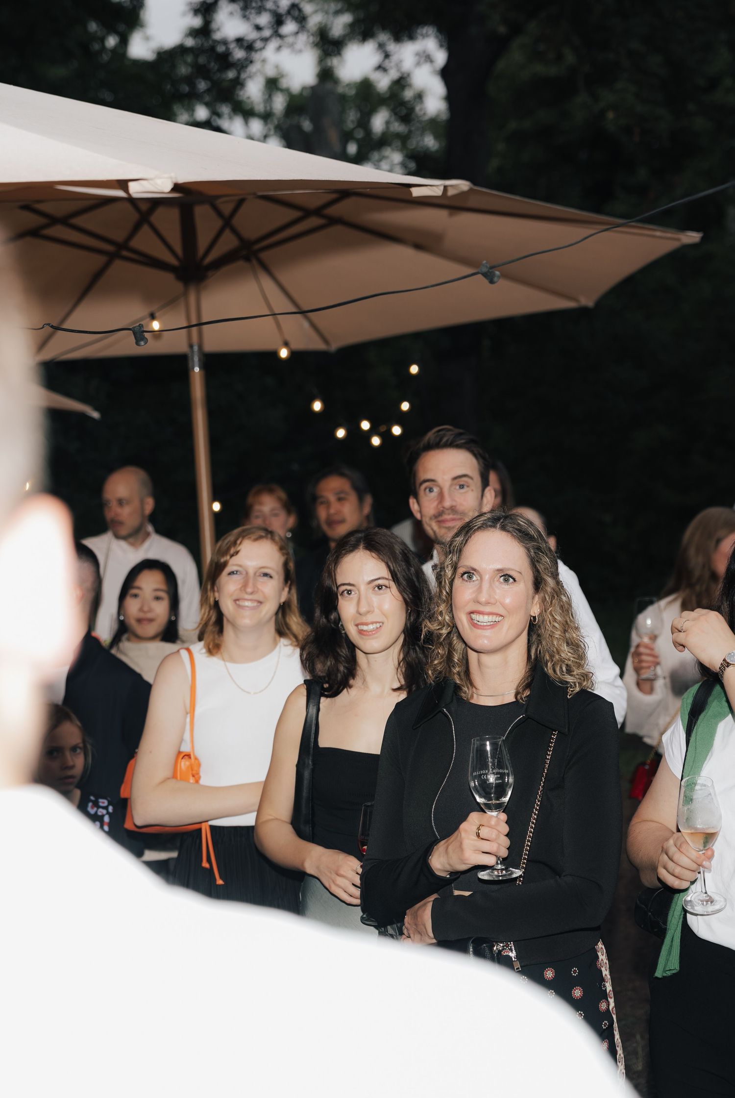 Group of guests mingling under white market umbrellas at an evening outdoor event.