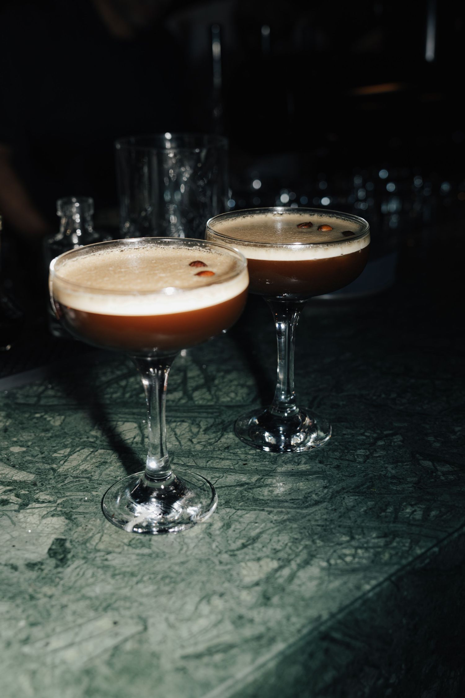 Cocktail glasses with frothy espresso martinis on a dark textured surface in moody lighting.