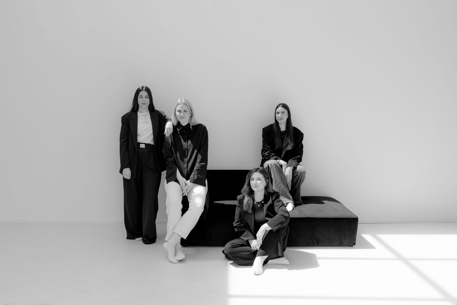 Four women in black outfits pose elegantly in a minimalist studio setting in a black and white photo.
