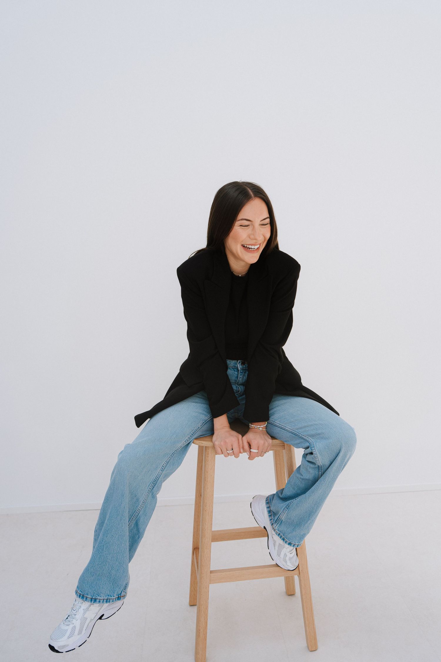 Smiling woman in black blazer and blue jeans sitting on wooden stool against white background.