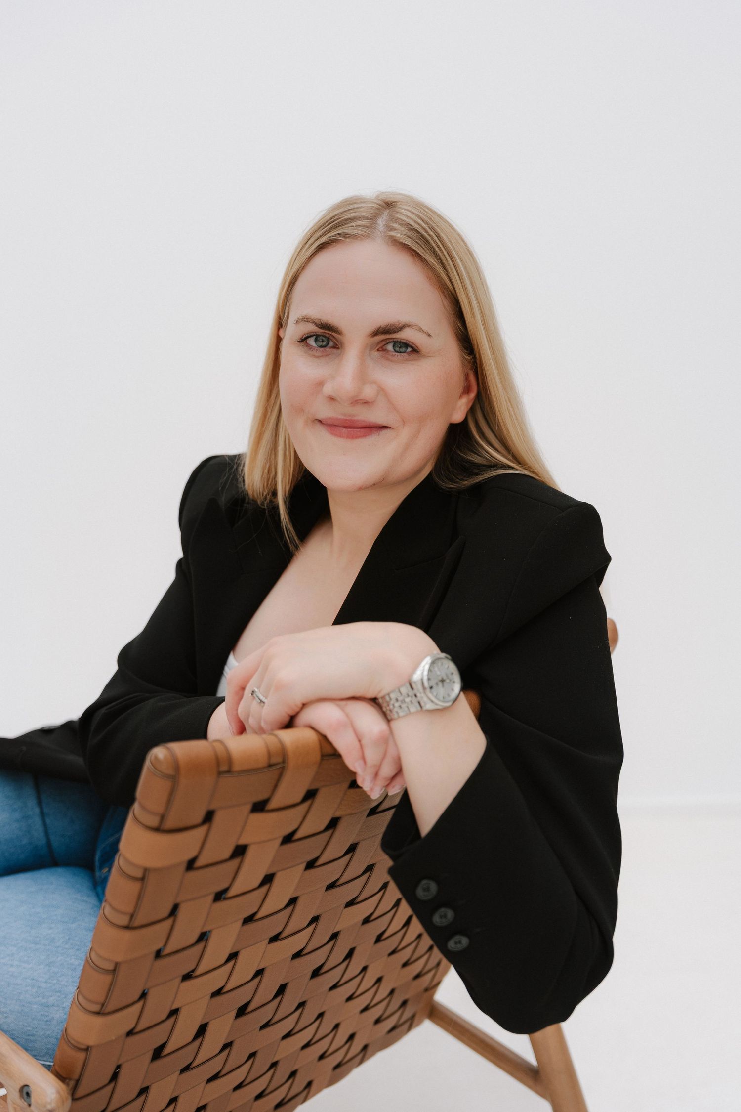 Woman in black blazer sitting relaxed on woven chair, smiling confidently against white background.