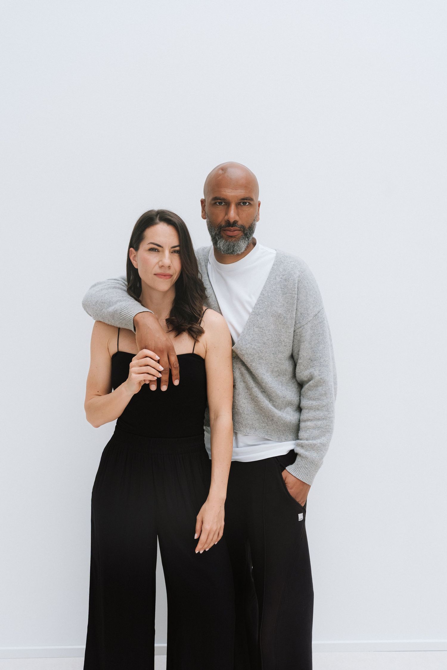 A man and woman pose together against a white background, she wears black and he wears a gray sweater.