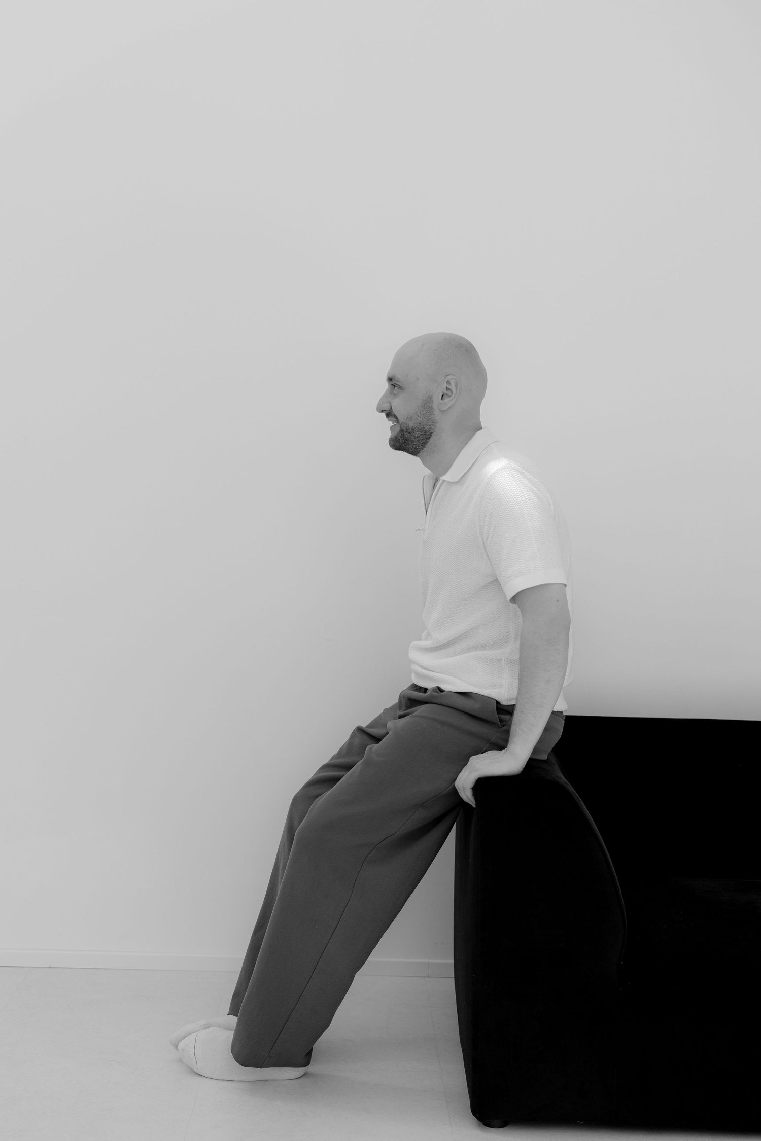 Bald man with beard in white polo and gray trousers leaning against a black sofa in a minimalist white room.