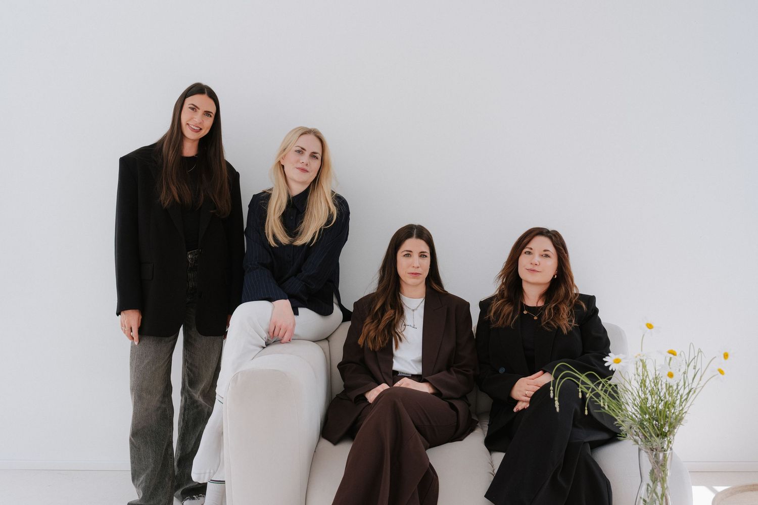 Four women in elegant dark outfits pose together in a minimalist white studio setting with a floral arrangement nearby.