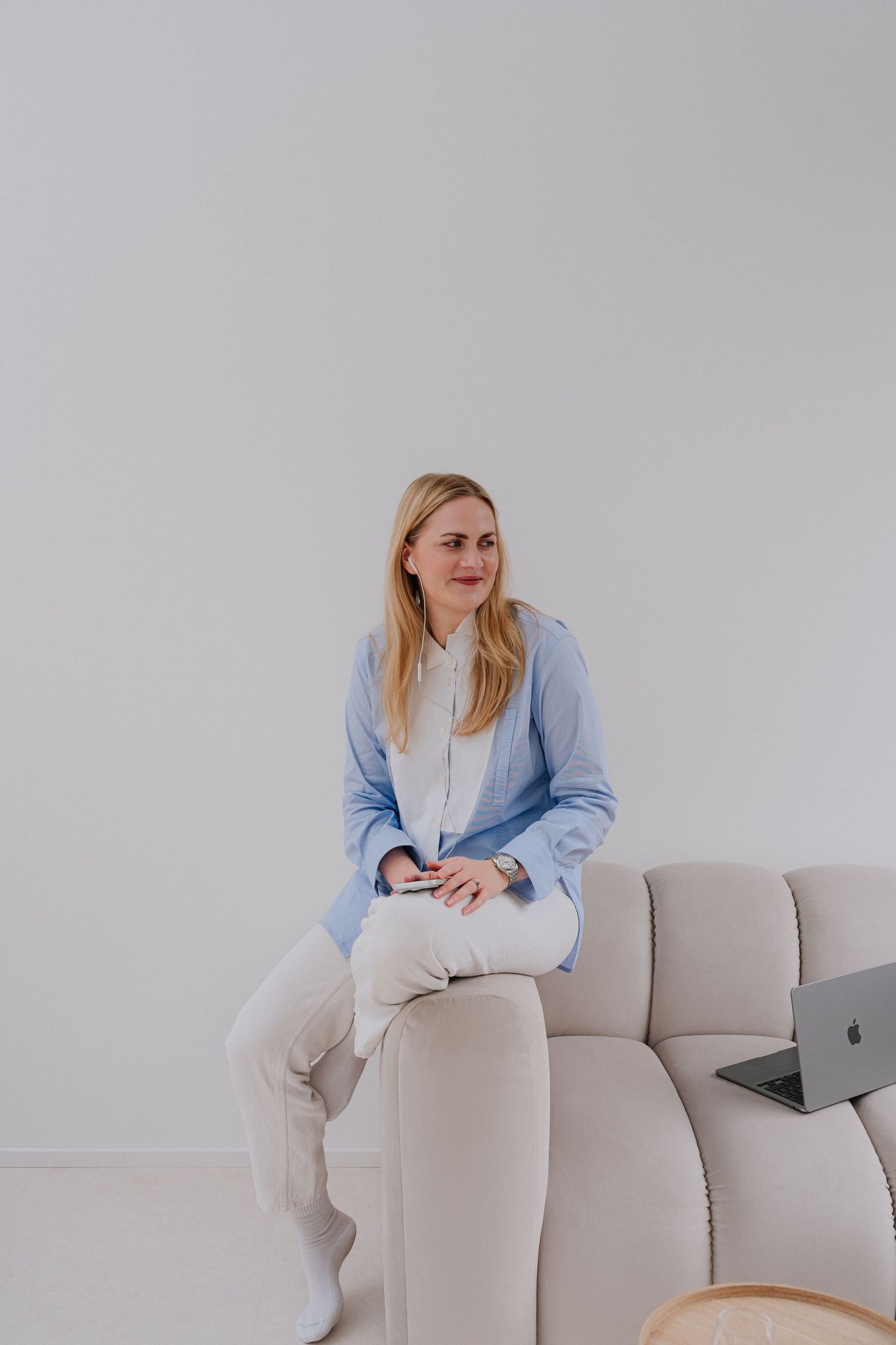 Blonde woman in light blue shirt and white pants sitting on beige sofa with laptop in bright minimalist room.