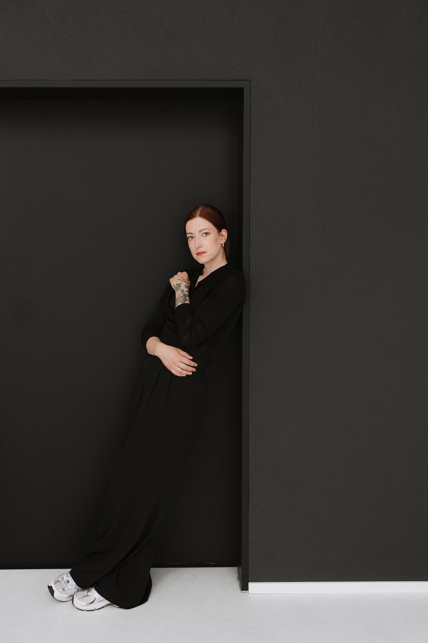 Woman in black outfit leaning against dark wall in minimalist setting, wearing white sneakers and tattoo sleeve.