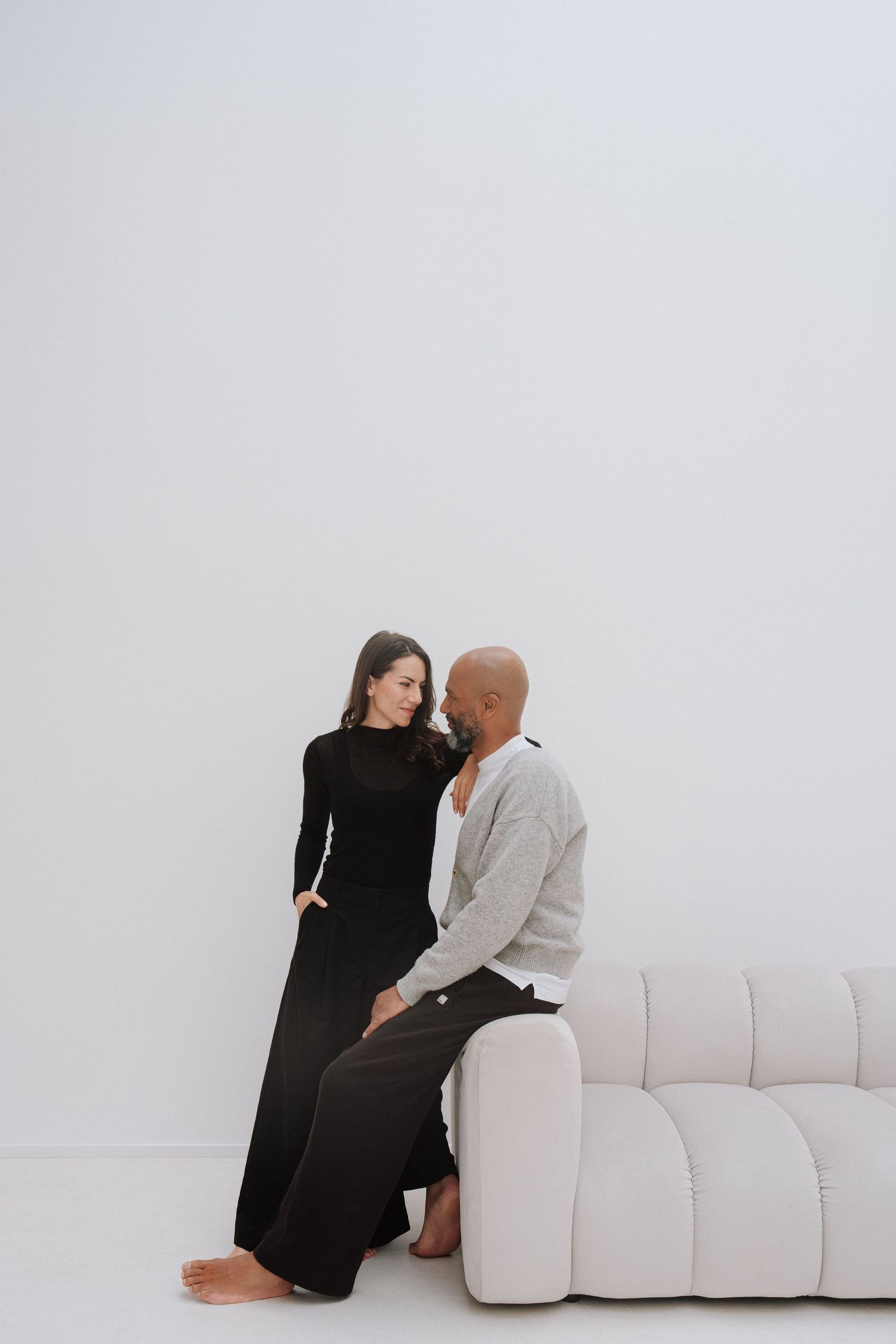 A couple in a minimalist white studio, woman in black dress and barefoot man in gray sitting on a white sofa.
