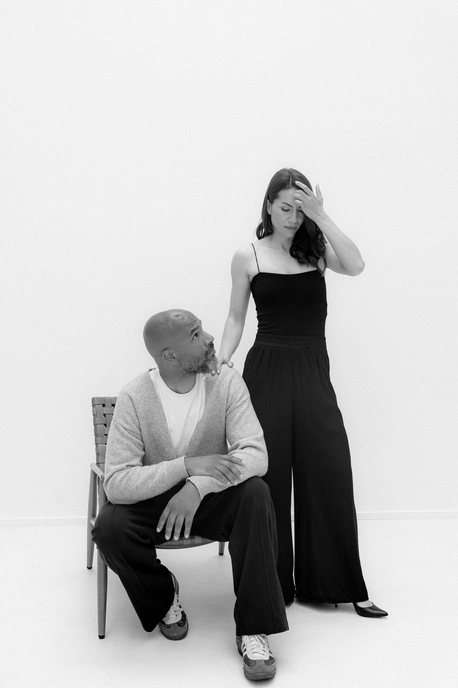 Black and white photo of a bald man sitting on a chair and a woman in black jumpsuit standing beside him.