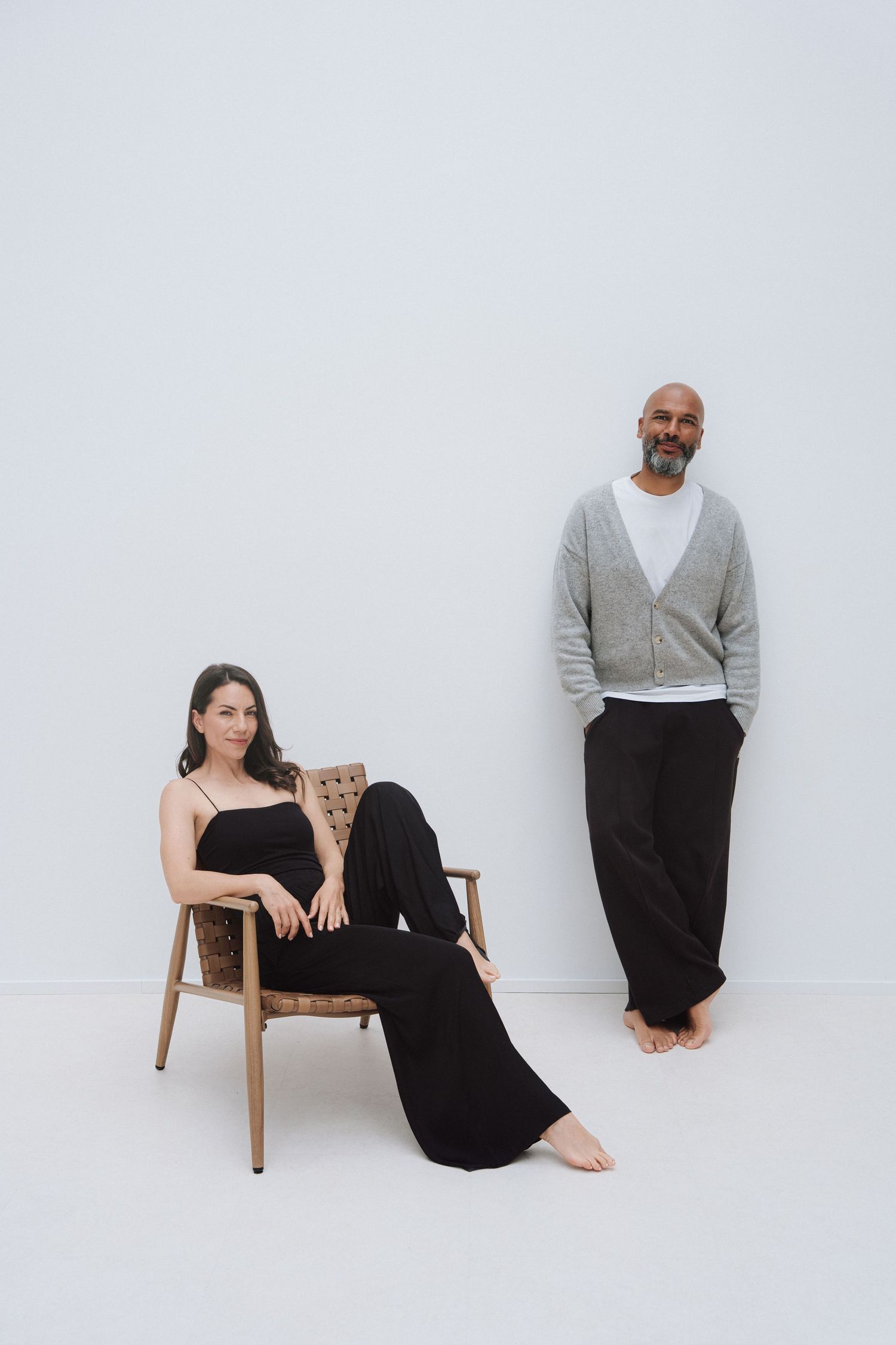 A woman in black sits in a wooden chair beside a standing man in grey cardigan and black pants against white wall.