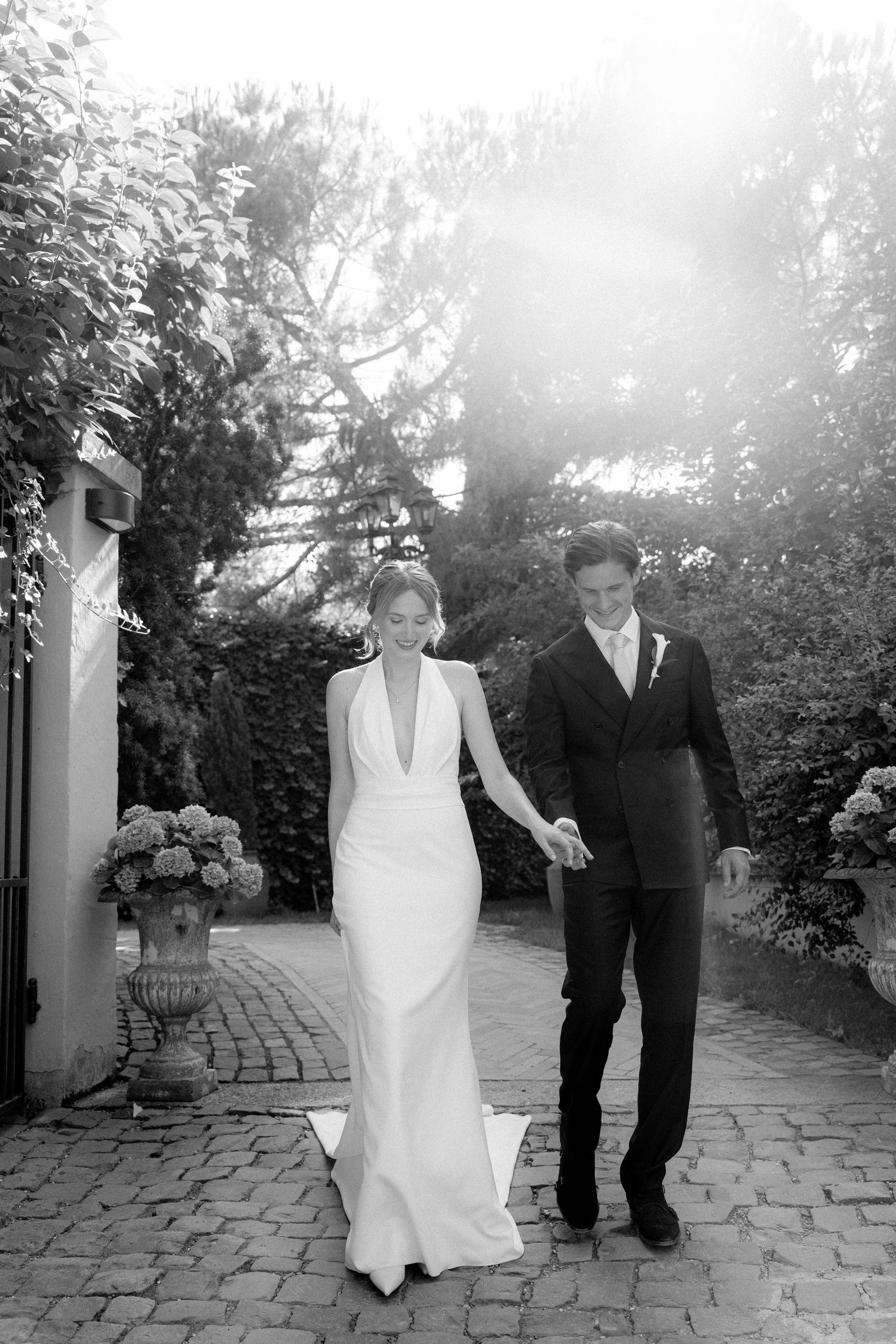 Black and white photo of a smiling bride and groom holding hands, walking through a sunlit garden on their wedding day.