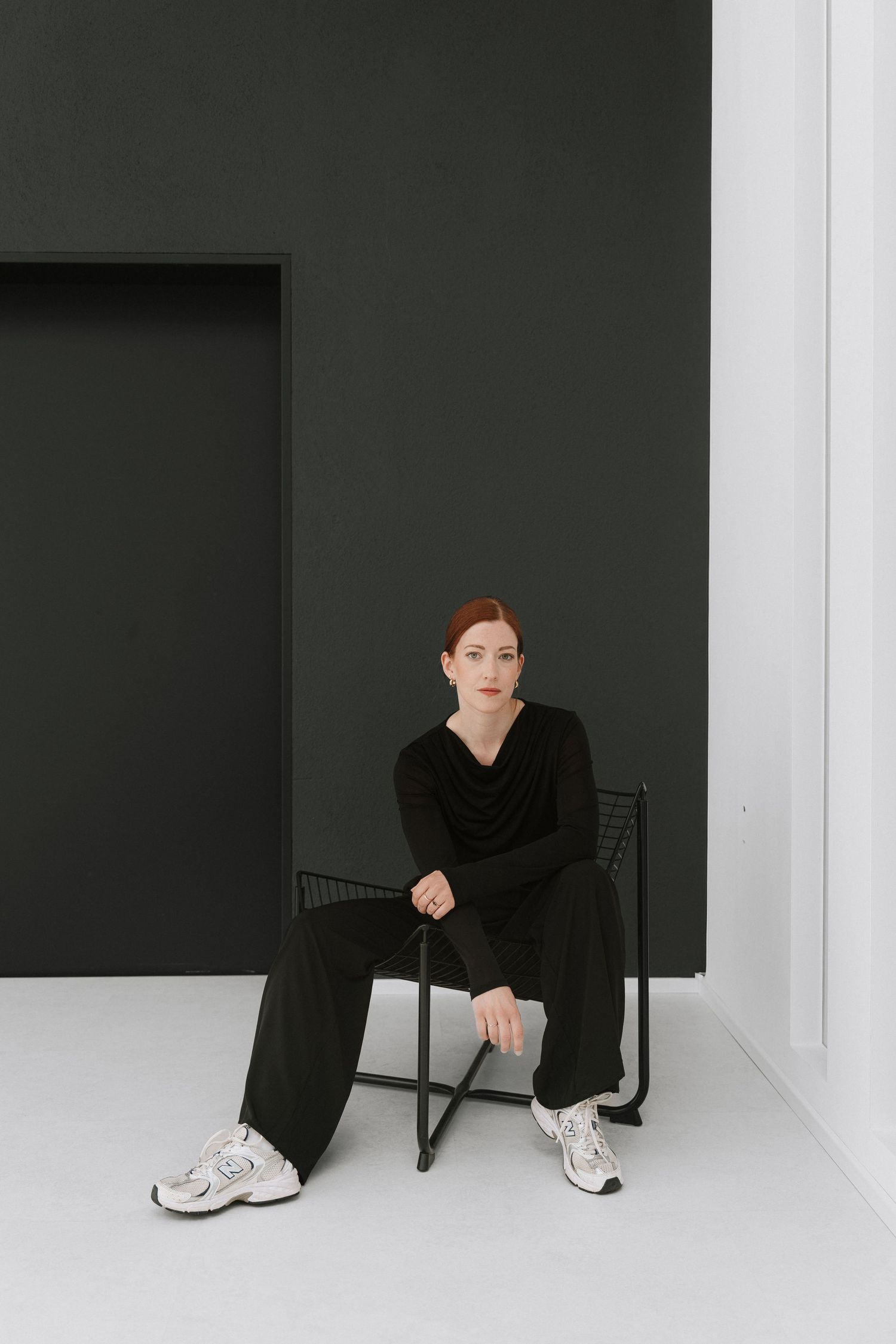 Woman in black outfit and white sneakers sitting on a metal chair against a dark green wall.