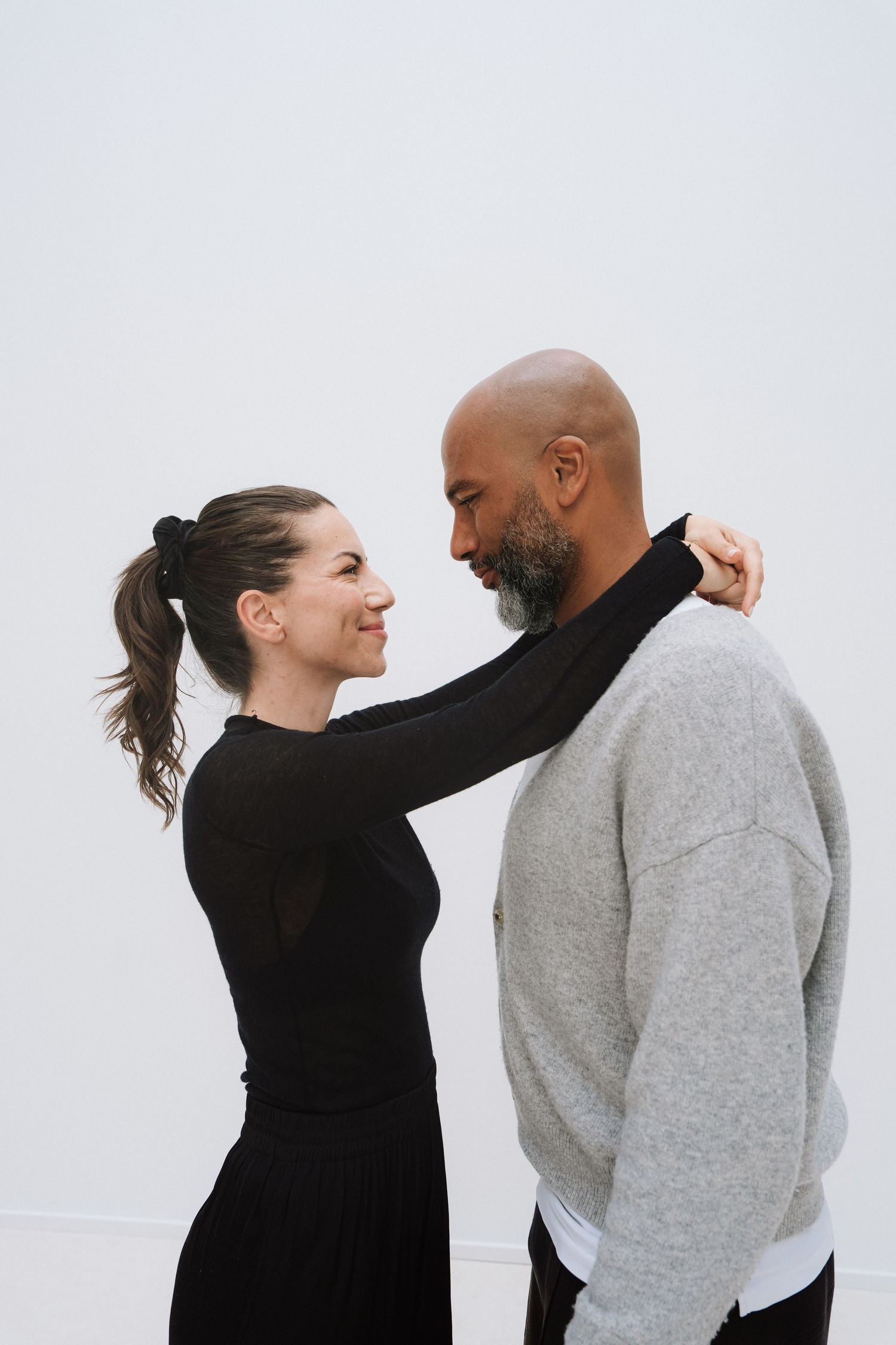 A smiling interracial couple sharing a tender moment, woman in black dress with hands on man's shoulders.