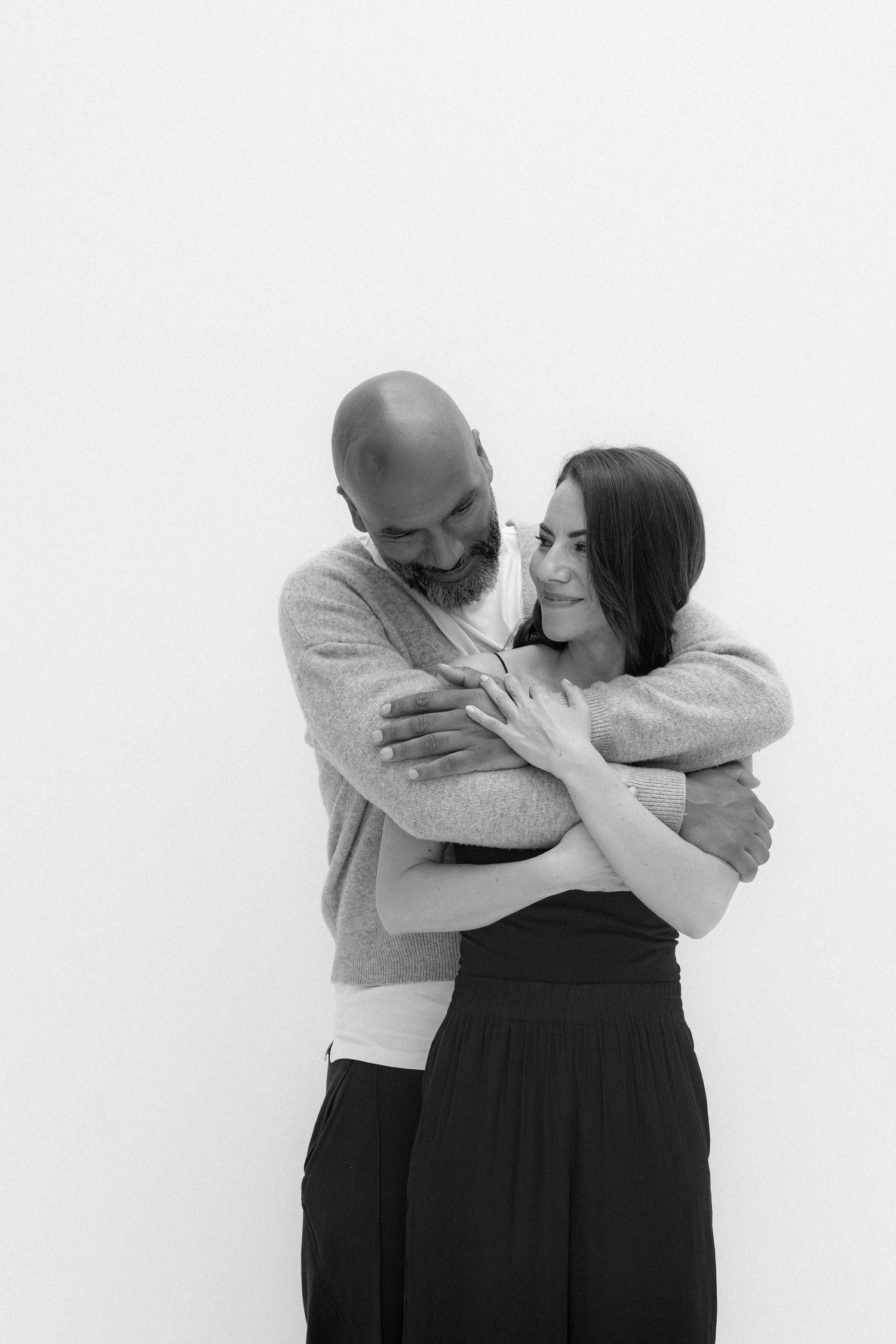 Black and white photo of a bald bearded man embracing a smiling dark-haired woman against a white background.