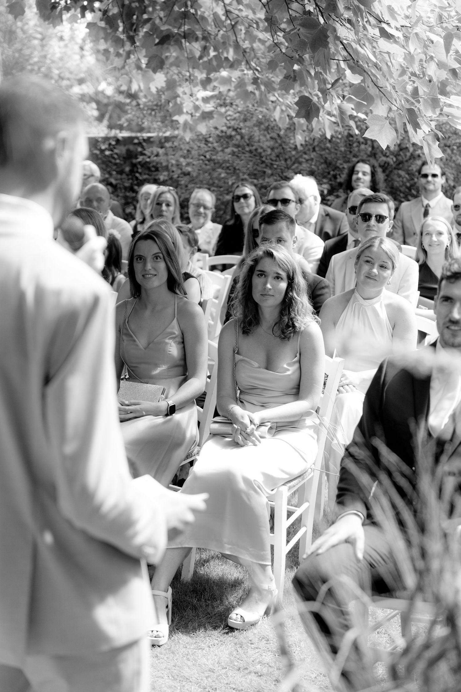 Black and white photo of wedding guests seated on white chairs during an outdoor ceremony.