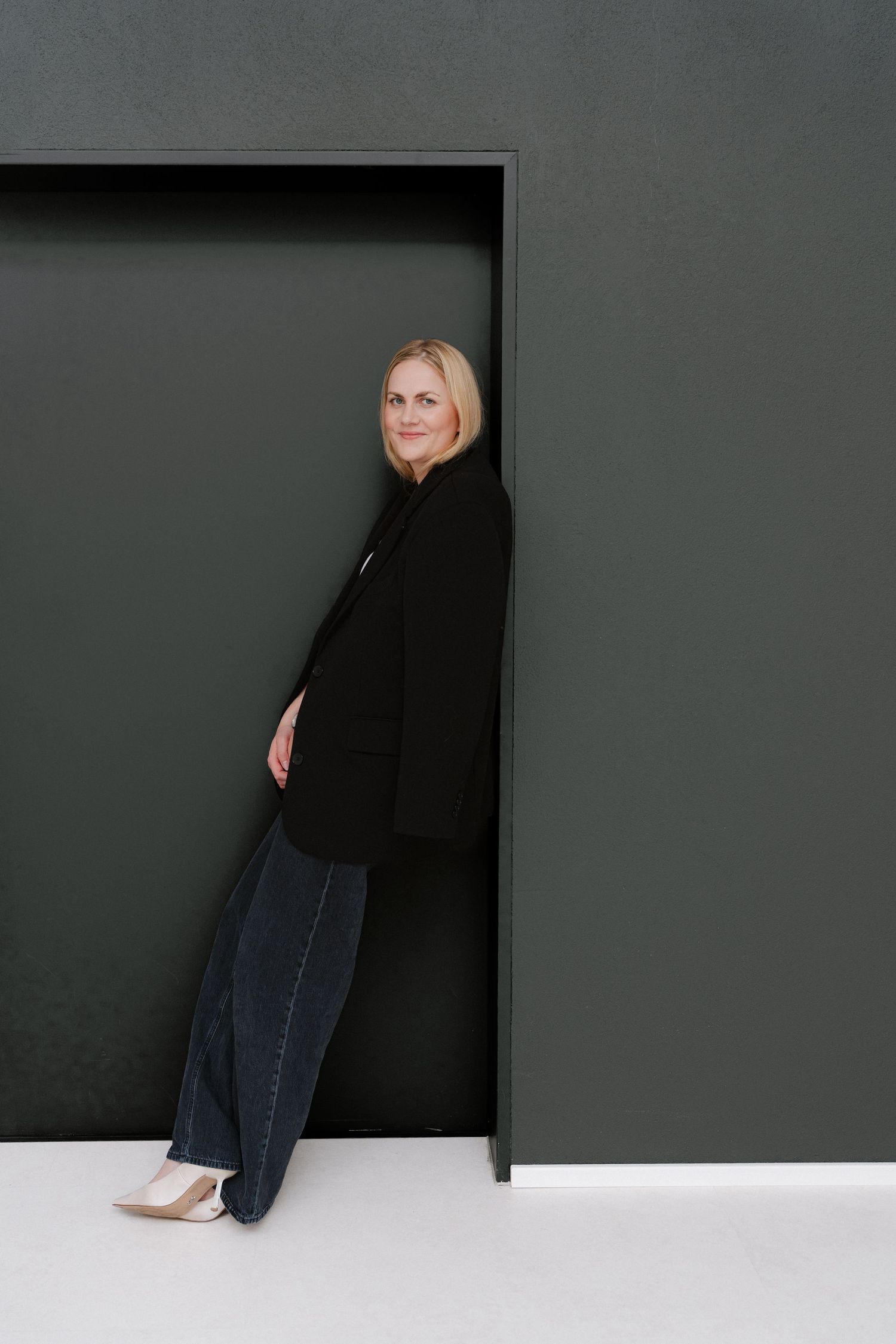 Blonde woman in black blazer and wide-leg pants leaning against a dark green wall, smiling confidently.