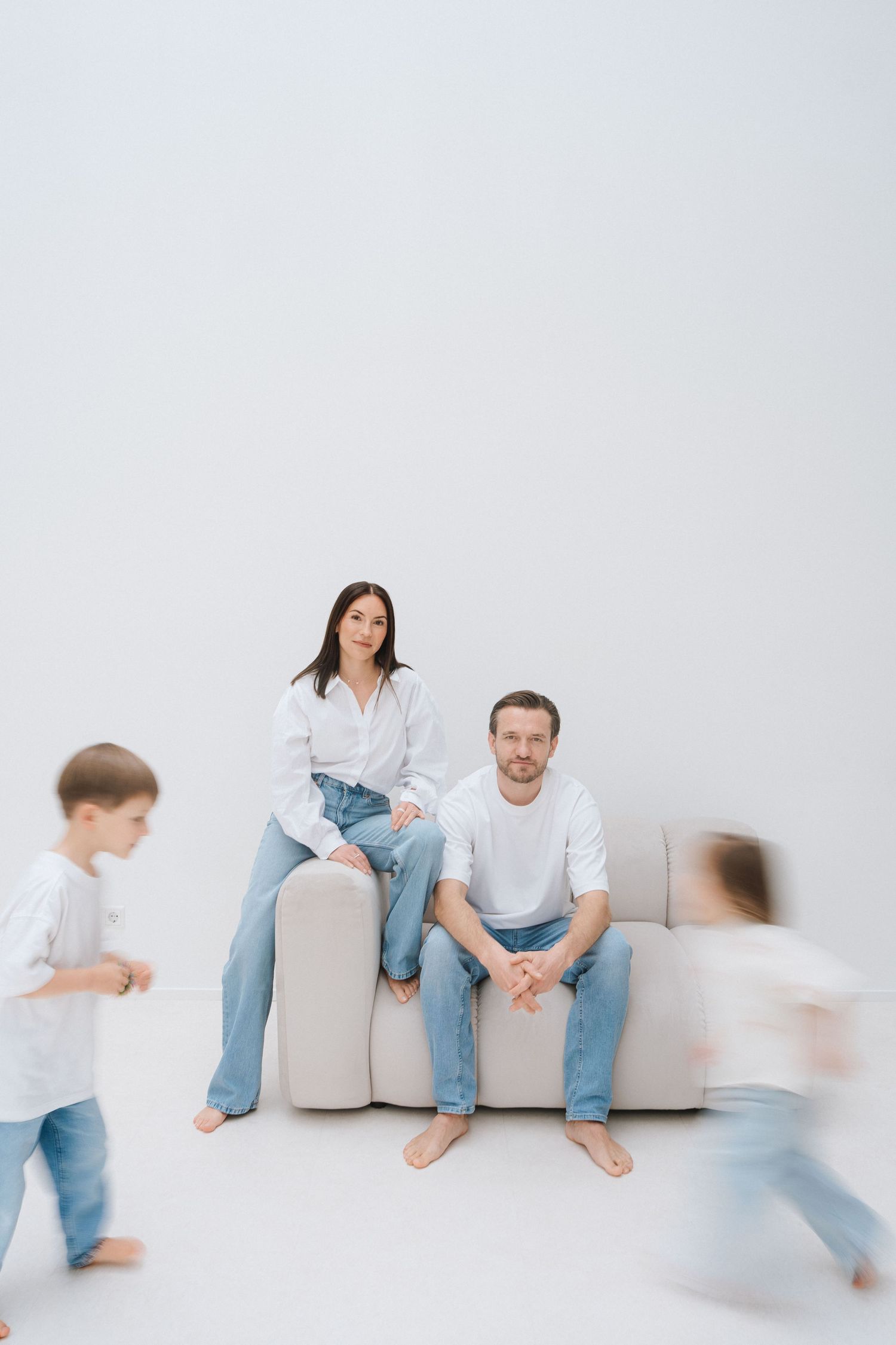 Family portrait with parents sitting on sofa in white and jeans while two children run past in motion blur.